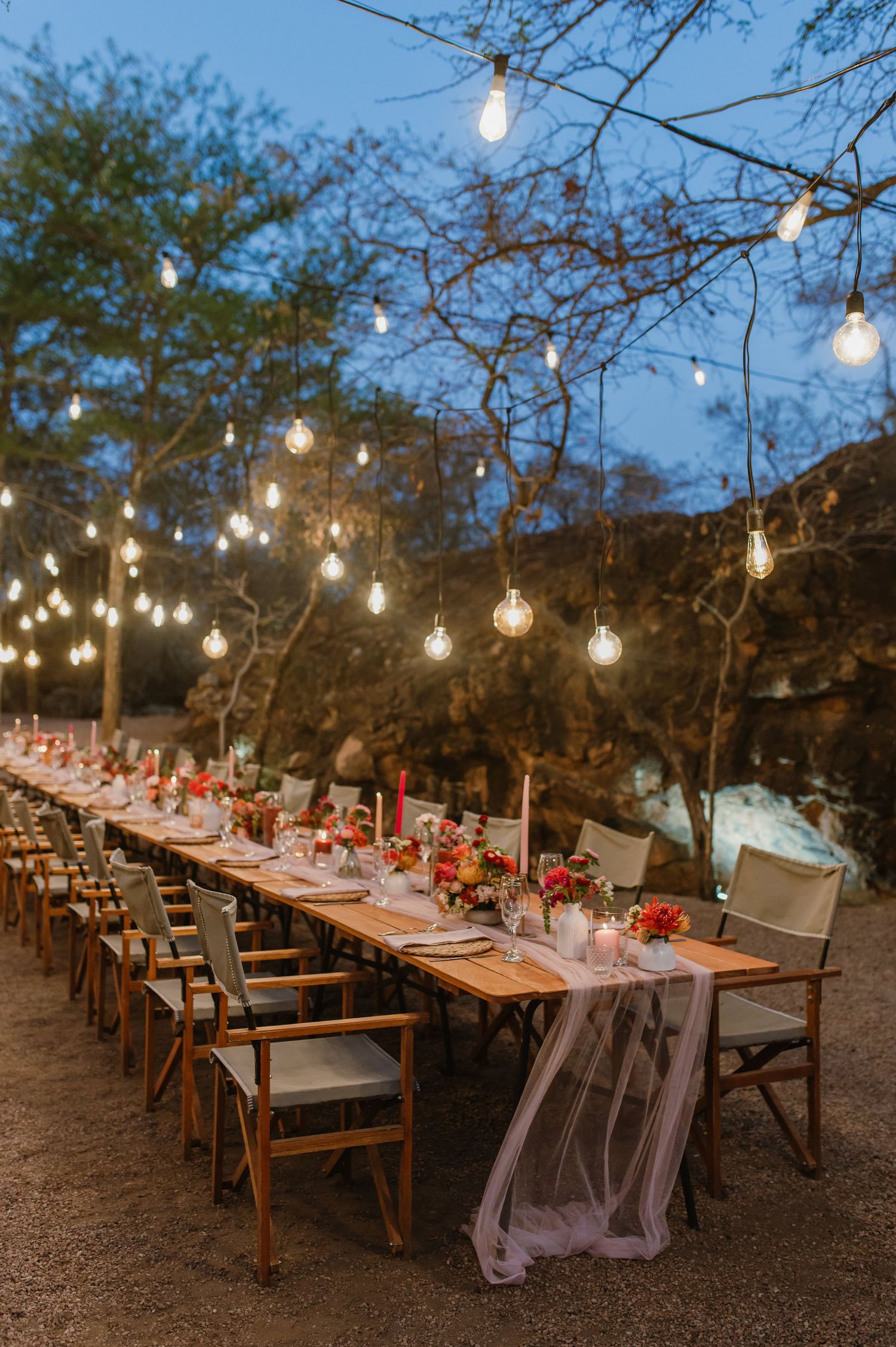 Long rustic dining tables set under string lights for an outdoor wedding reception.