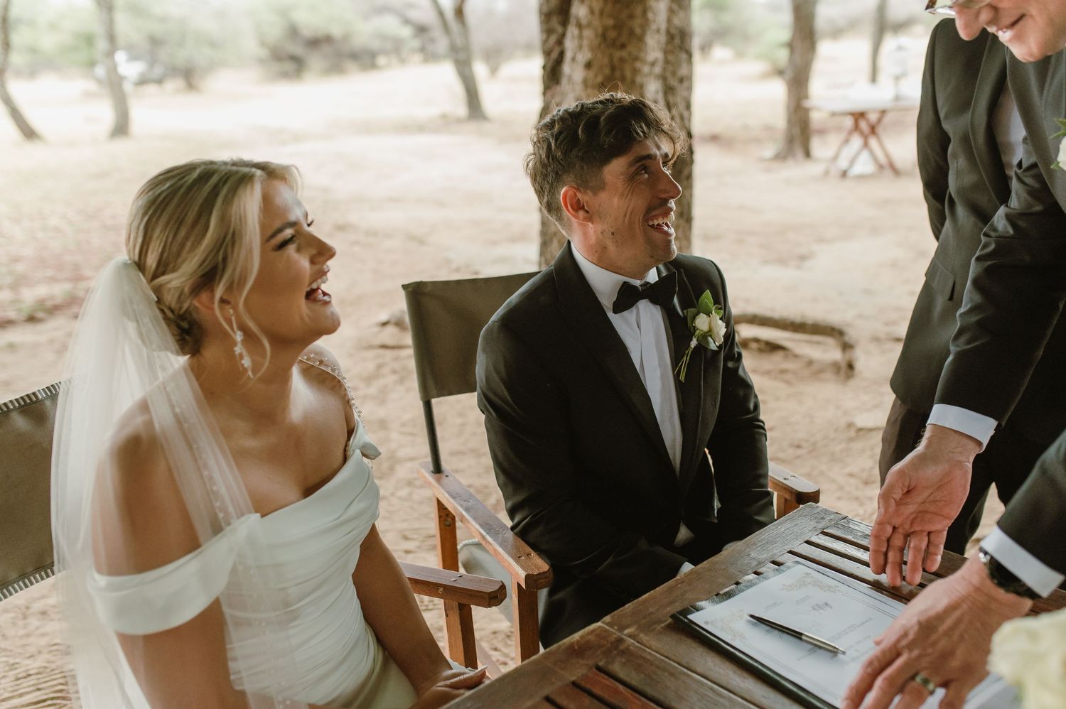 Wedding ceremony guests gather outdoors on wooden benches in a natural setting.