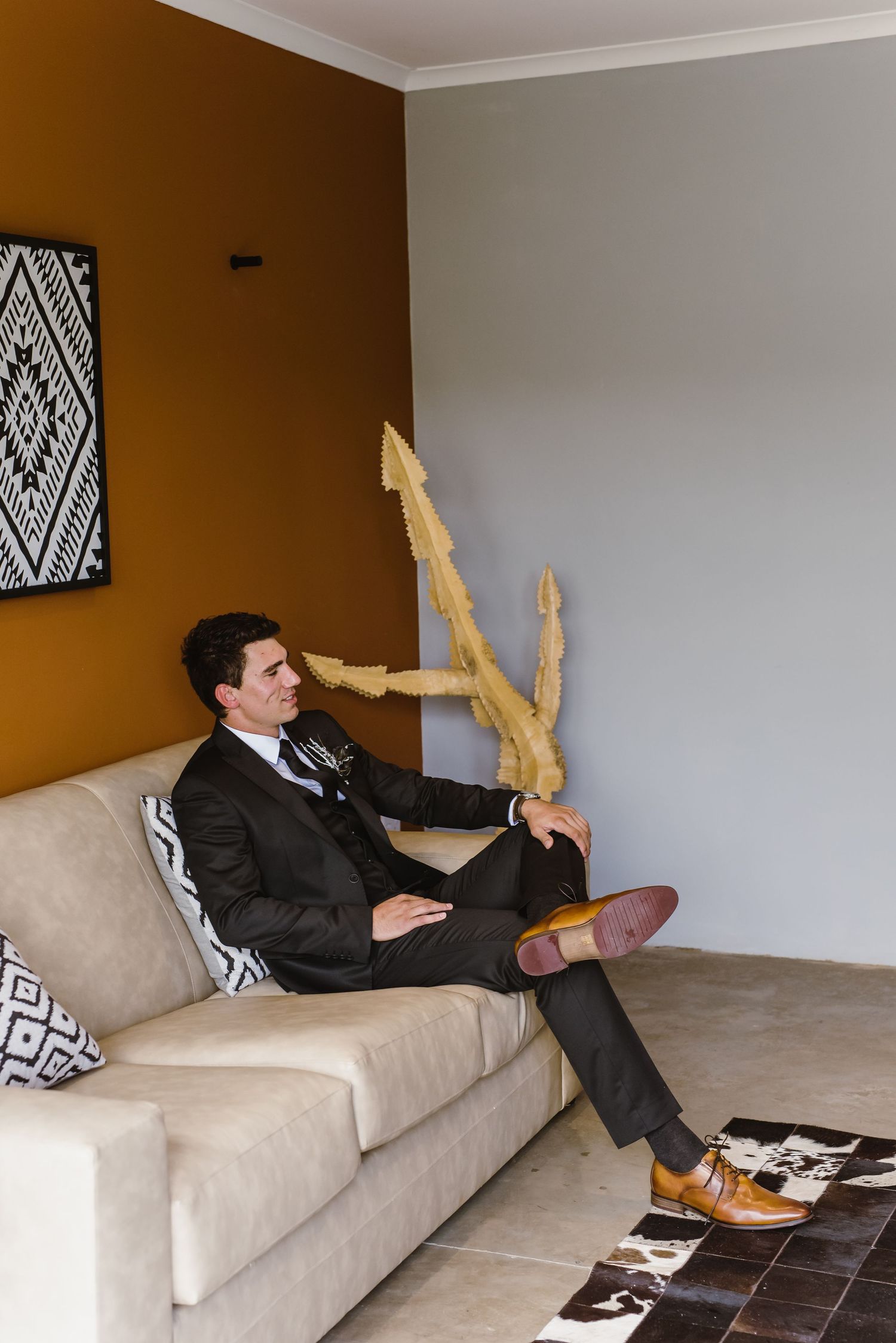 Groom relaxing on beige couch with feet up against geometric patterned wall.