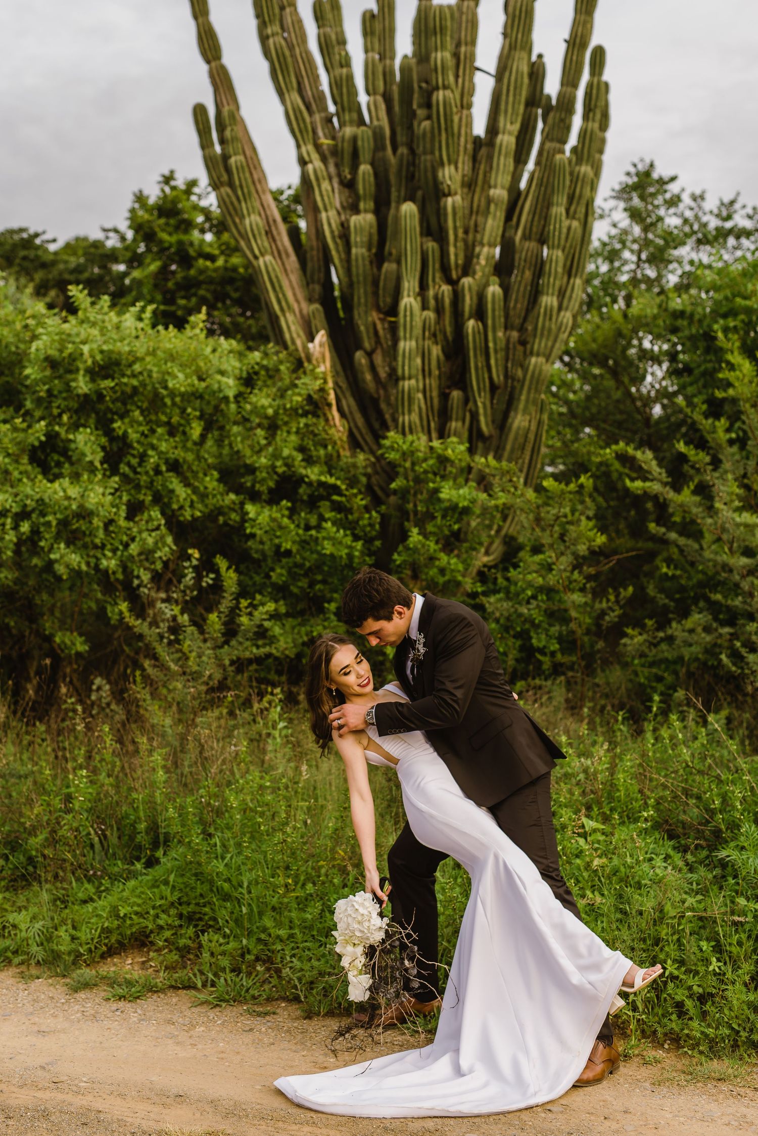 Artistic wedding pose with couple near tall cactus in desert garden setting.