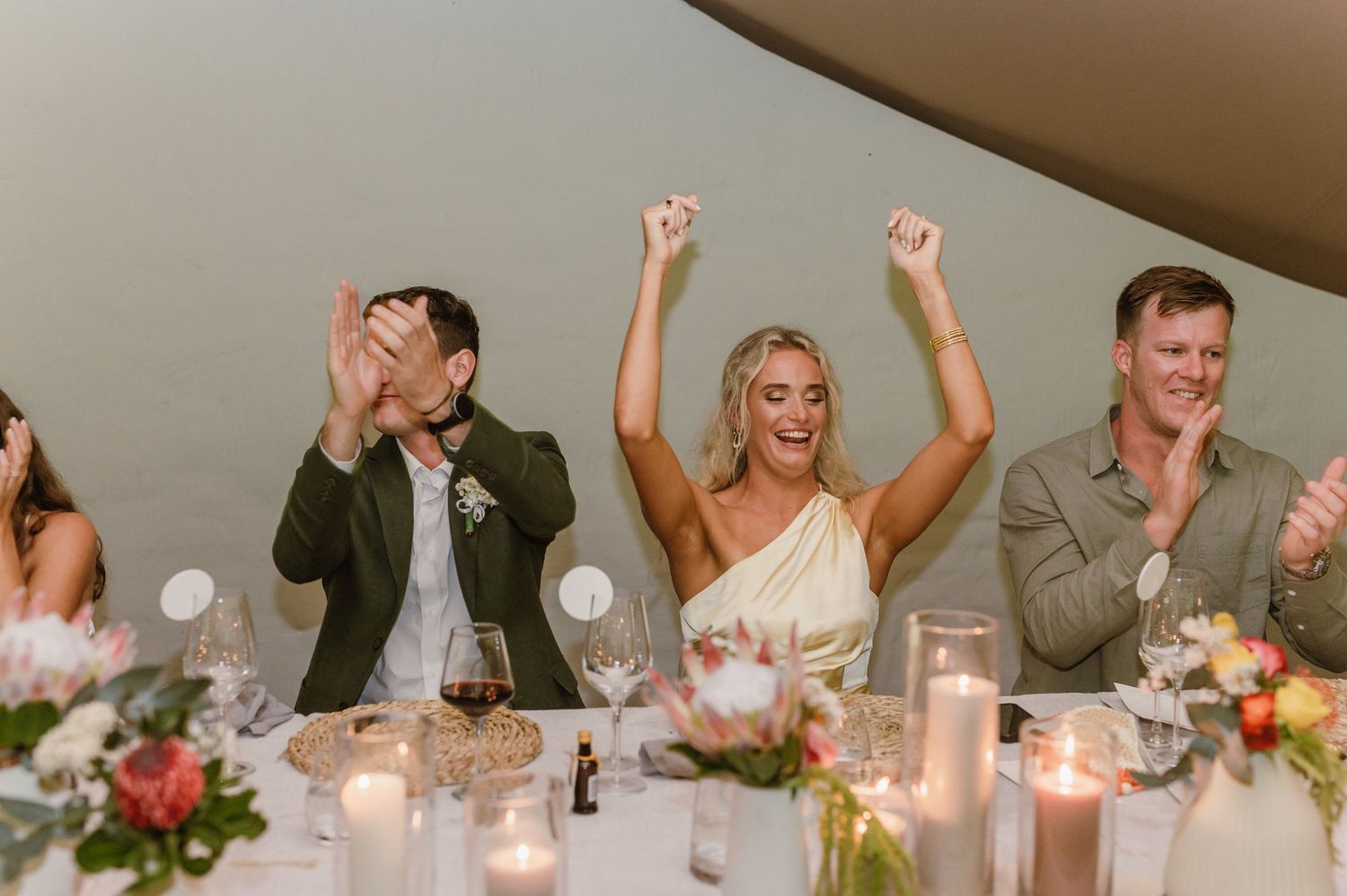 Guests celebrating and raising their hands during wedding toasts at a candlelit reception dinner.