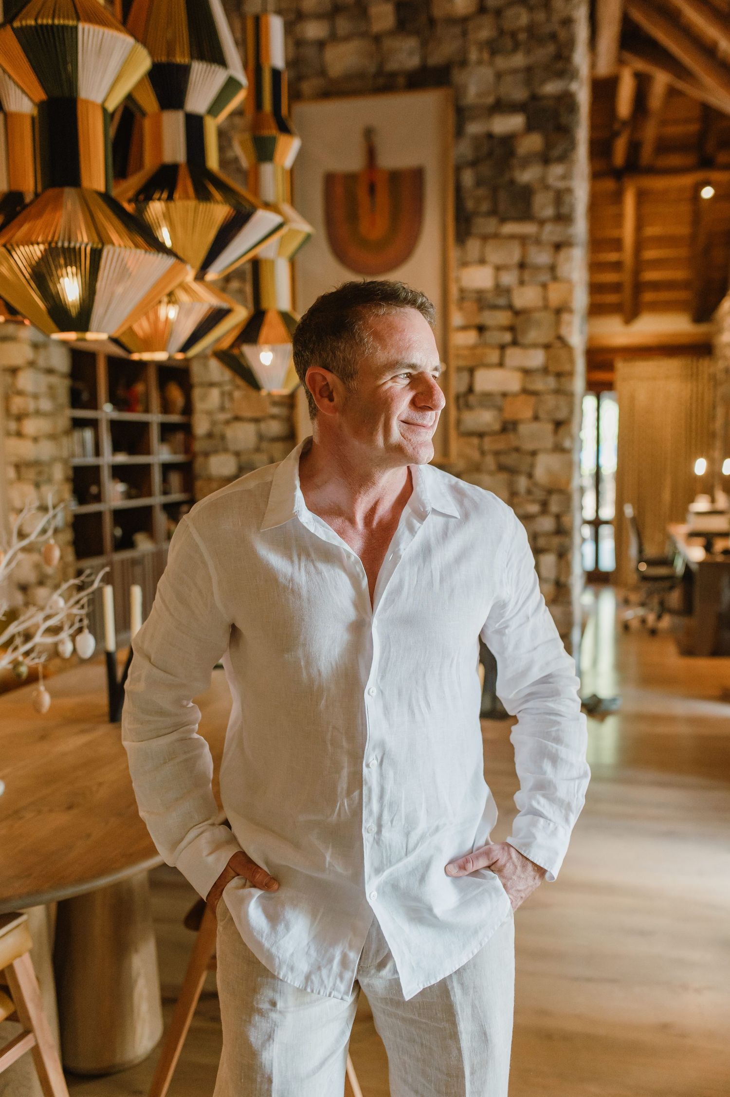 Man in white linen shirt and pants standing in rustic interior with wooden pendant lights.