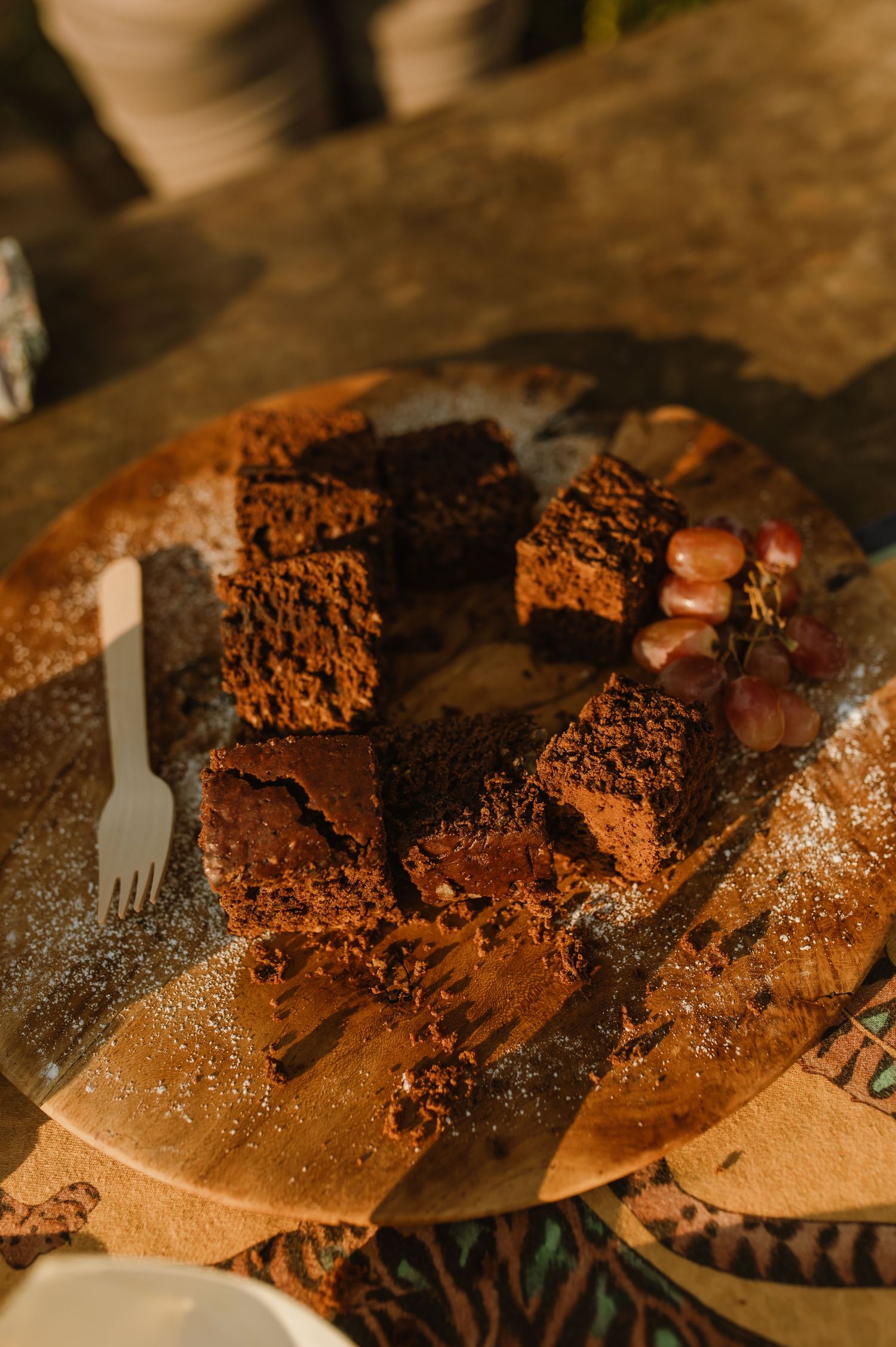 Chocolate brownies dusted with powdered sugar on a wooden board with a white plastic fork and fresh grapes.