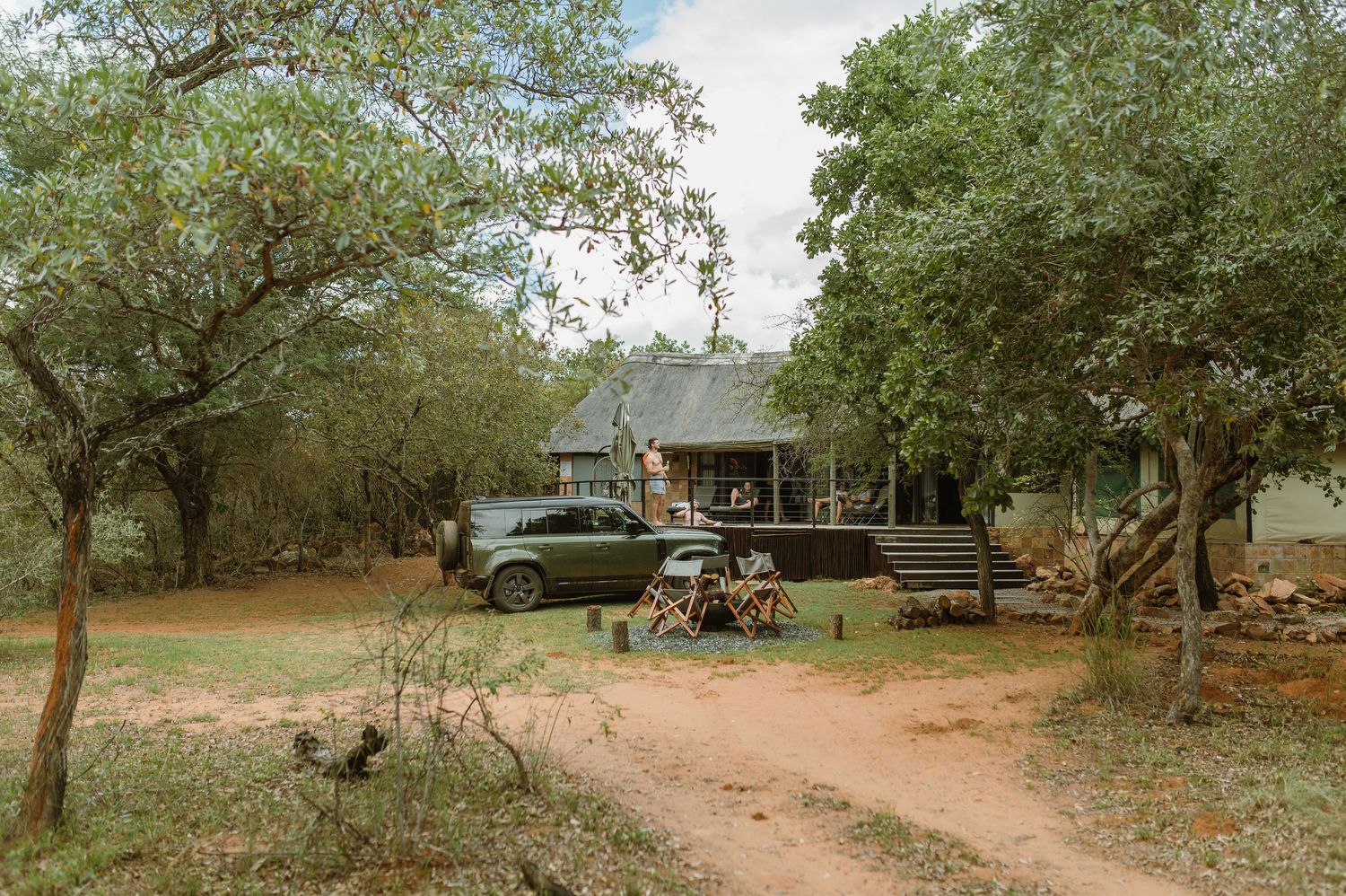 Safari lodge surrounded by trees with a parked SUV and rustic outdoor seating on a dirt path in a wildlife reserve.
