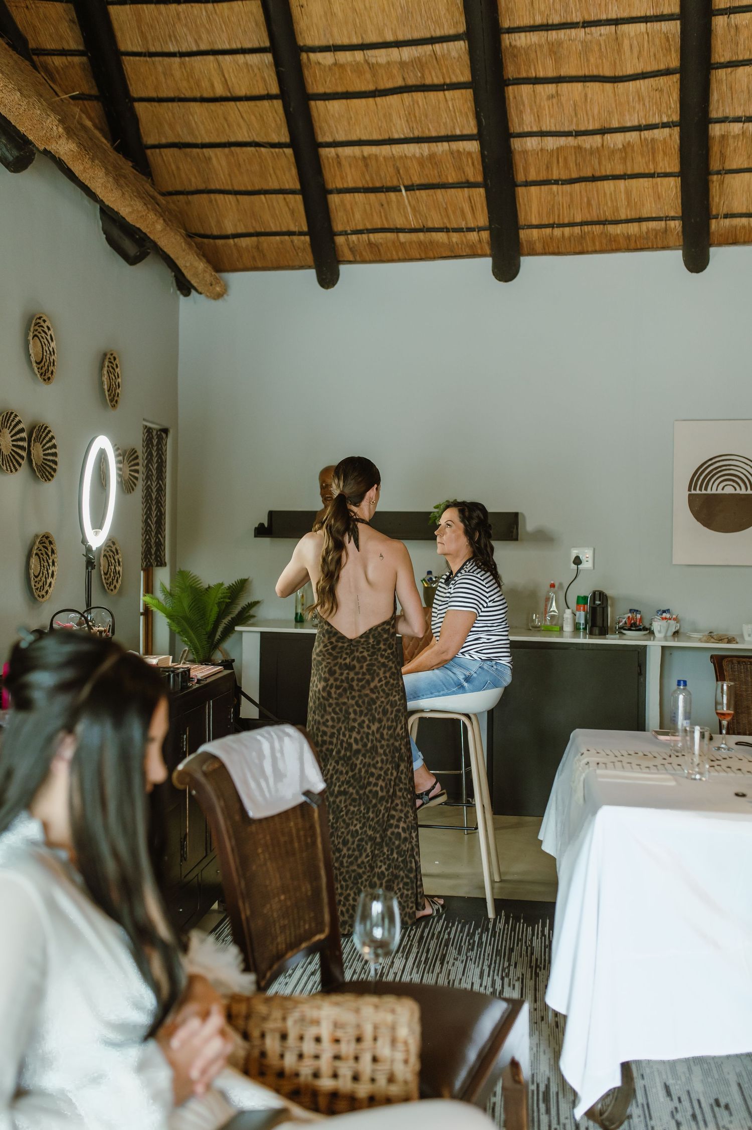 Women gather in a rustic thatched-roof room for bridal preparations with a makeup mirror and styling station.
