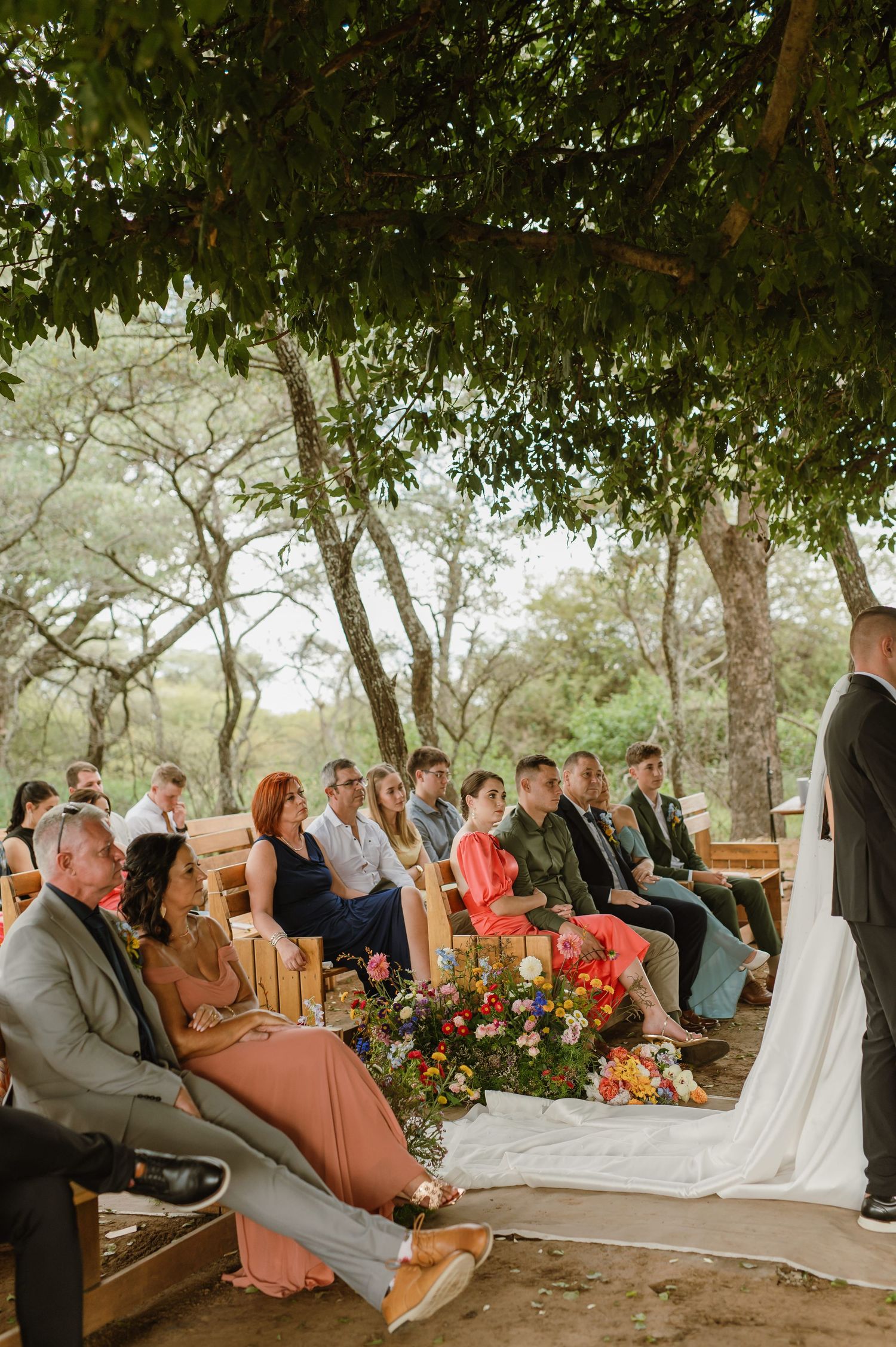 Outdoor wedding ceremony under trees with guests seated on wooden benches and colorful floral arrangements.