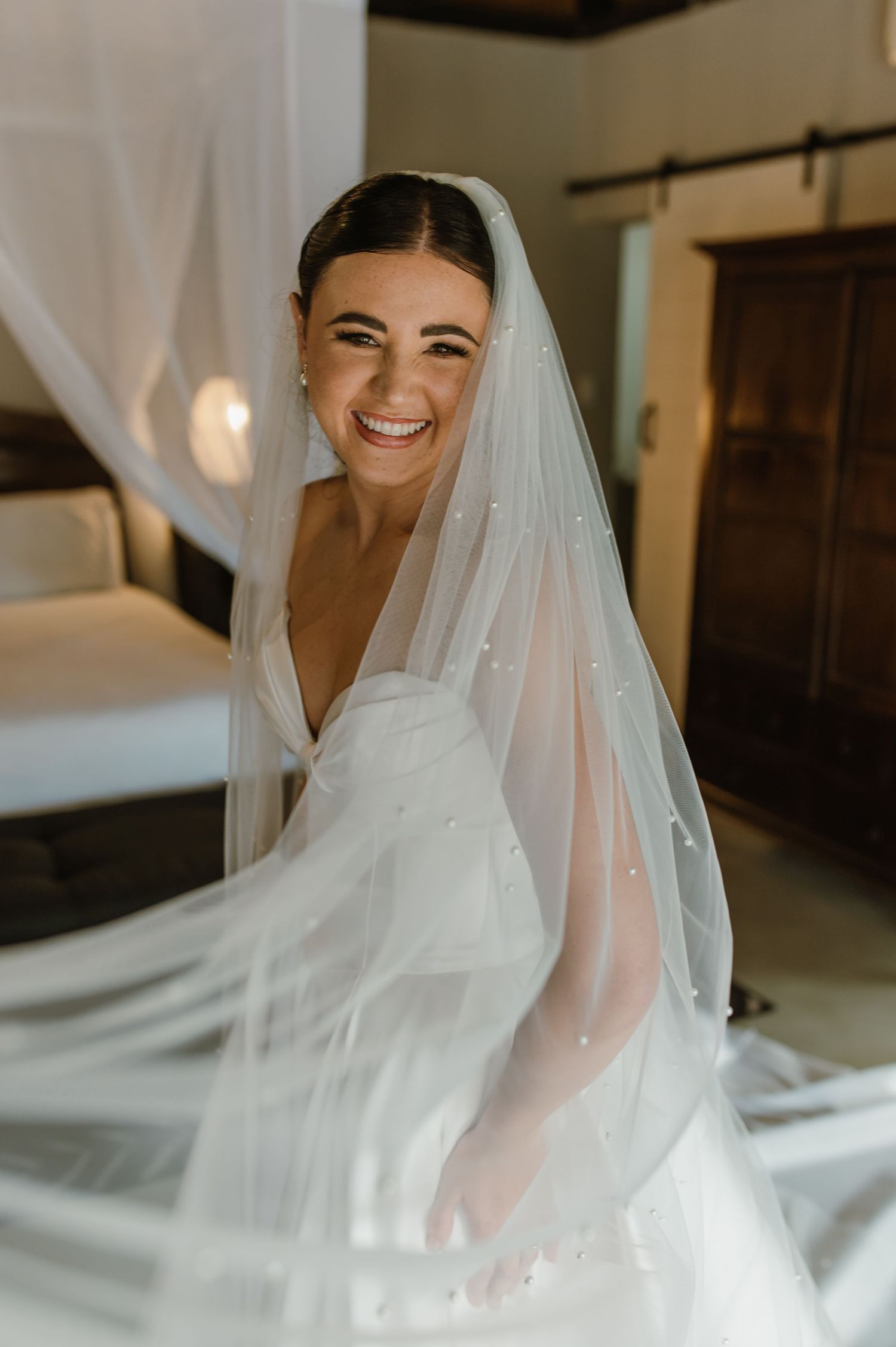 Joyful bride smiling through flowing pearl-embellished veil in a rustic wooden wedding suite.