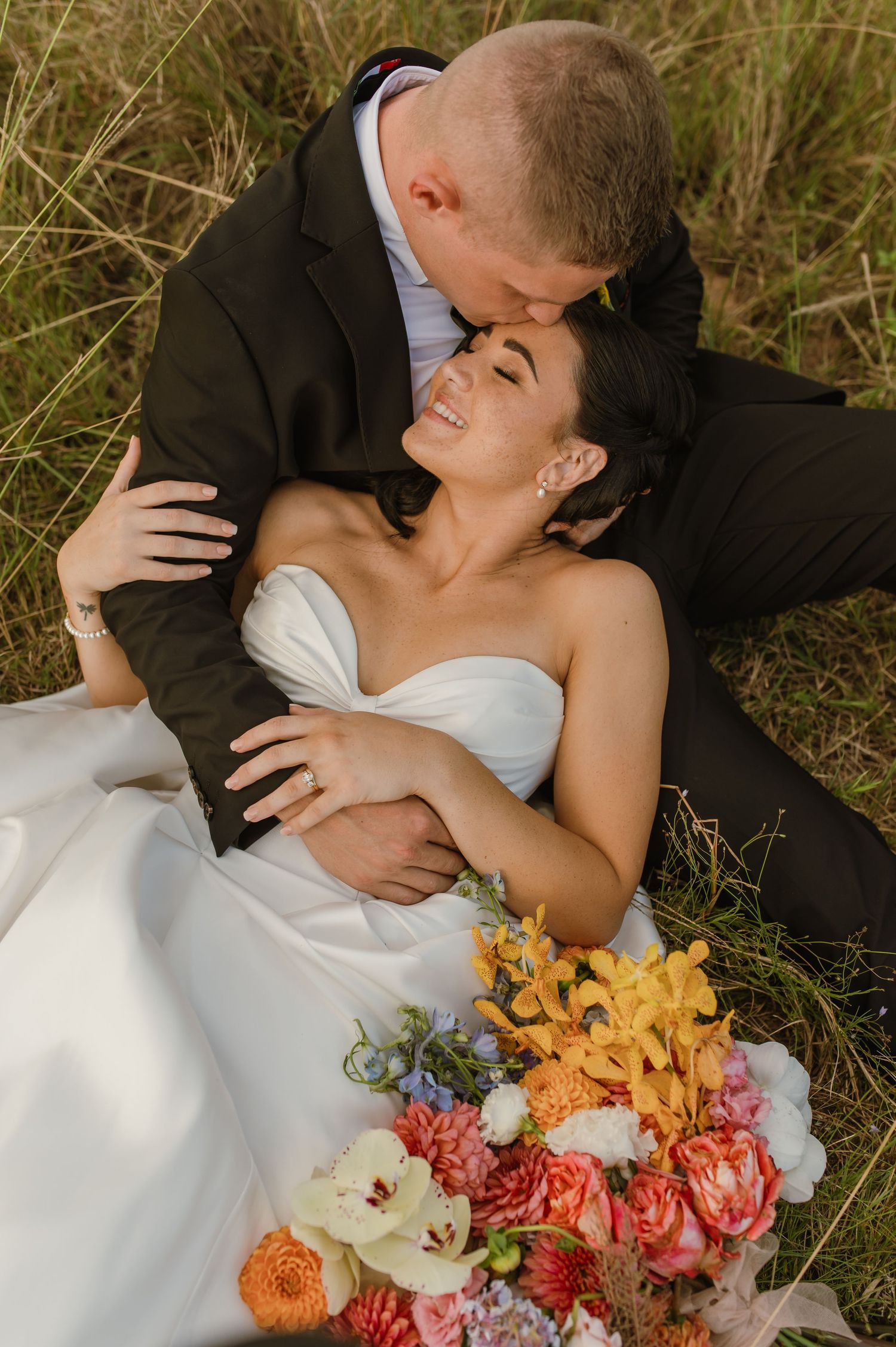 Groom kisses bride on forehead as she smiles, reclining in grass with vibrant floral bouquet nearby.