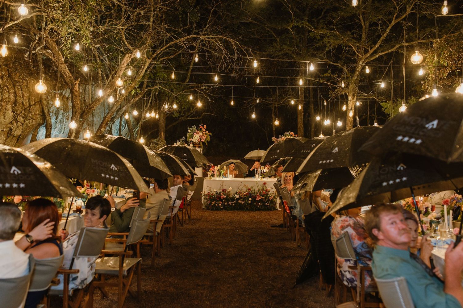 Outdoor wedding reception under string lights with guests holding black umbrellas in a festive atmosphere.