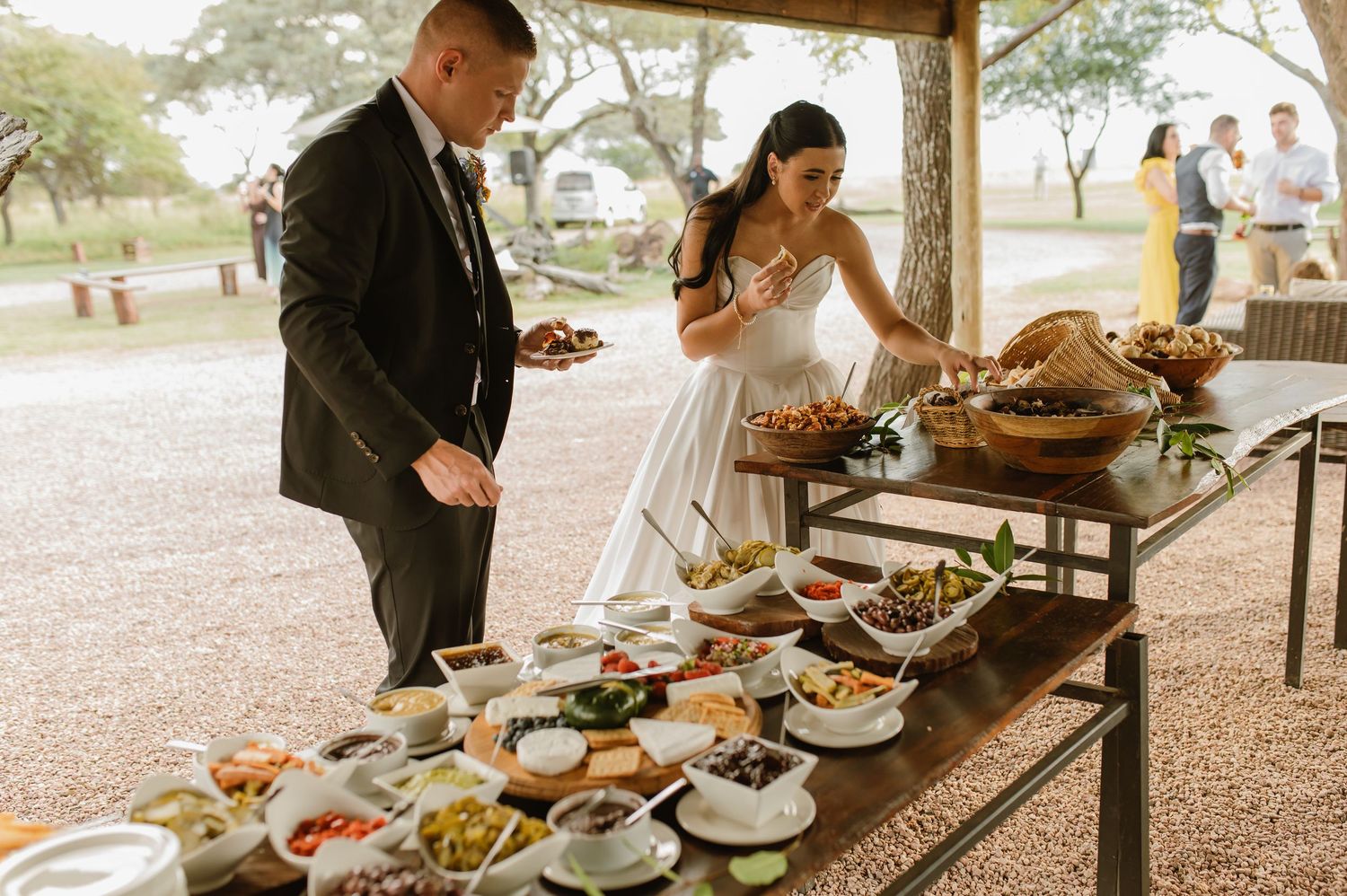Bride and groom serve themselves at elegant outdoor wedding buffet spread with variety of dishes.