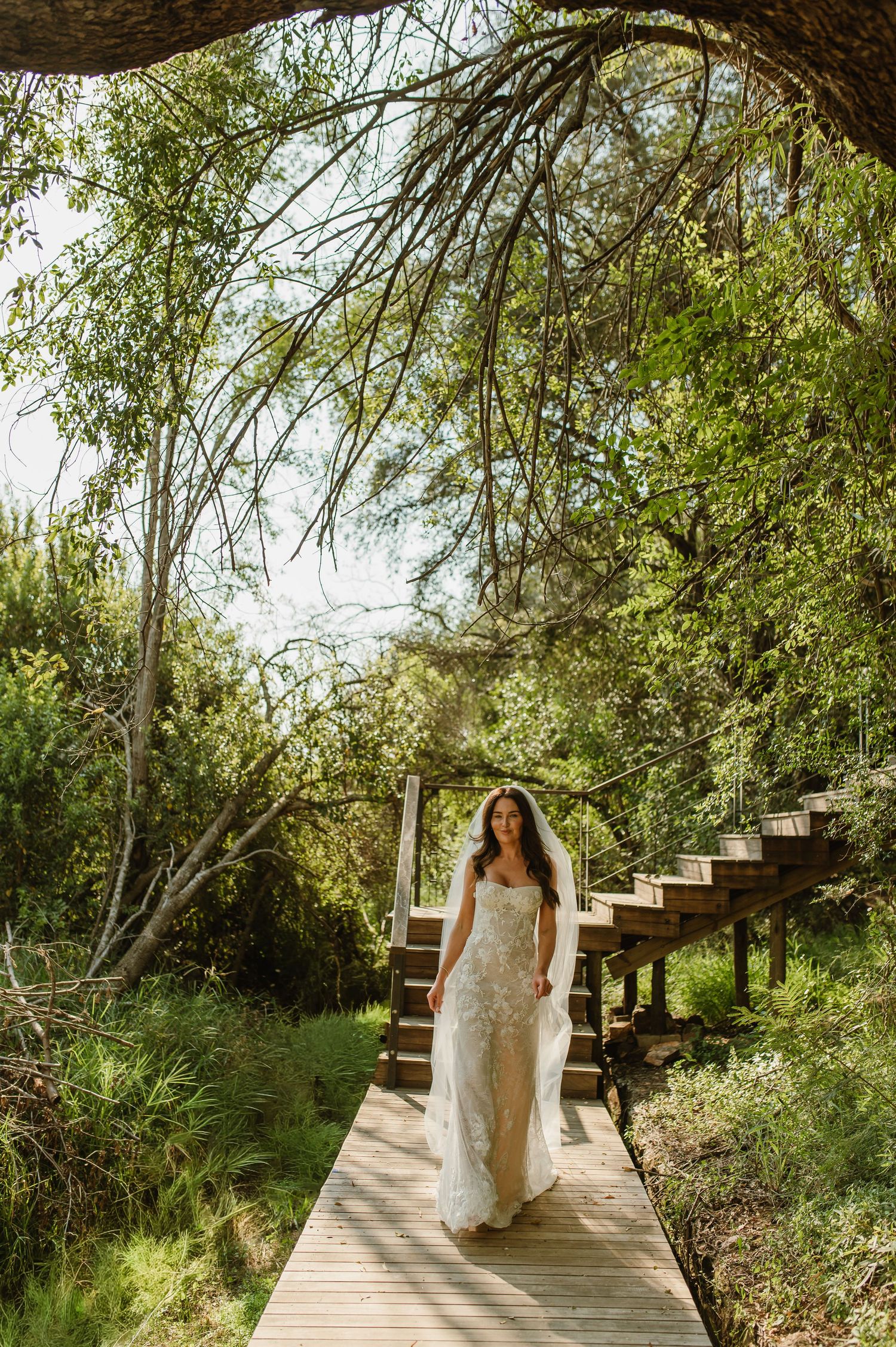 Bride in lace wedding dress and veil walking along wooden boardwalk path through lush green forest.