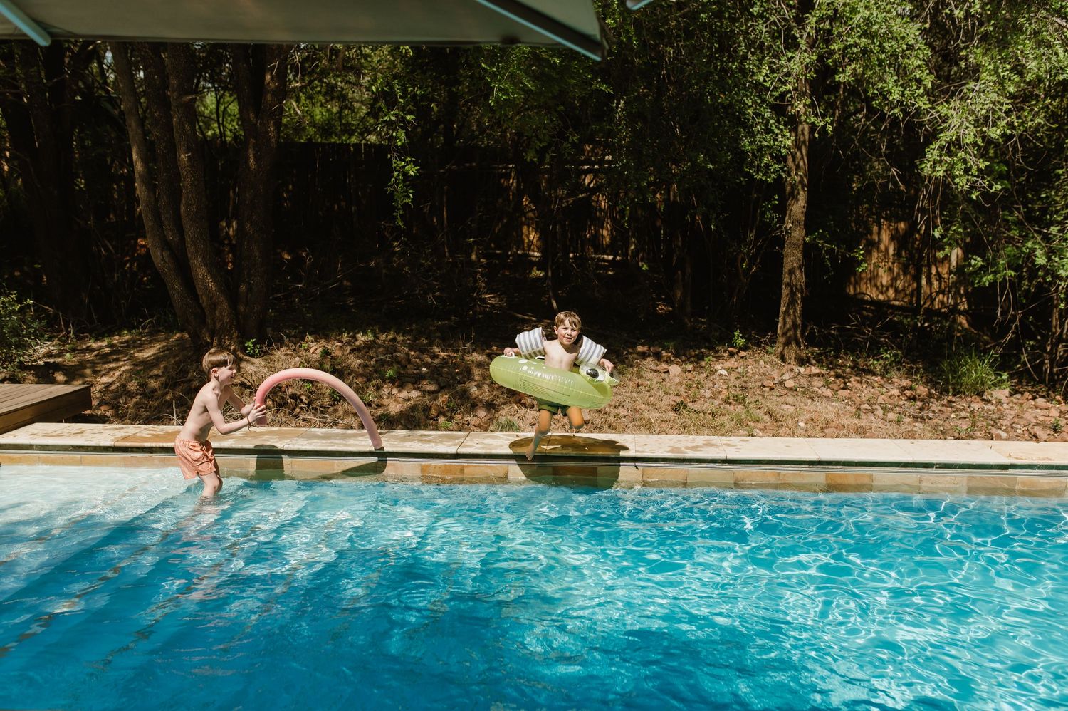 Two children playing in a sunny outdoor swimming pool surrounded by lush green trees.