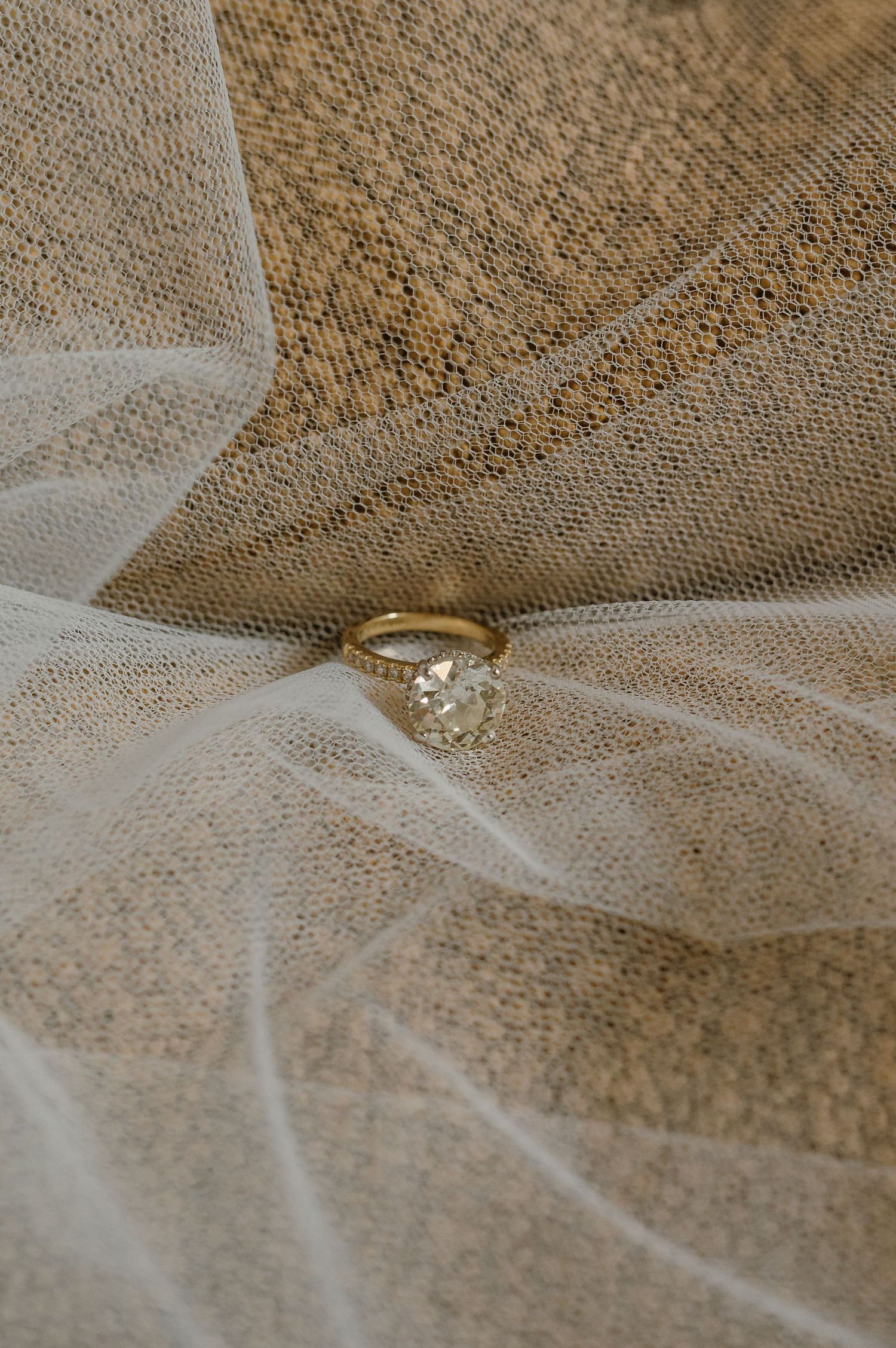 A gold solitaire diamond engagement ring resting on a sheer white veil and woven fabric.