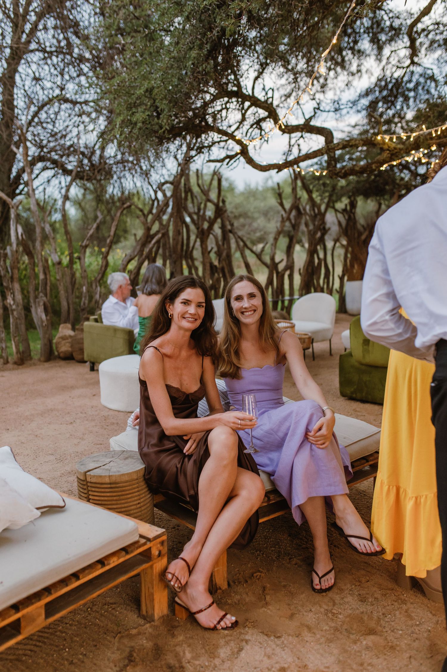 Guests enjoying an outdoor reception with rustic seating and string lights under trees at dusk.