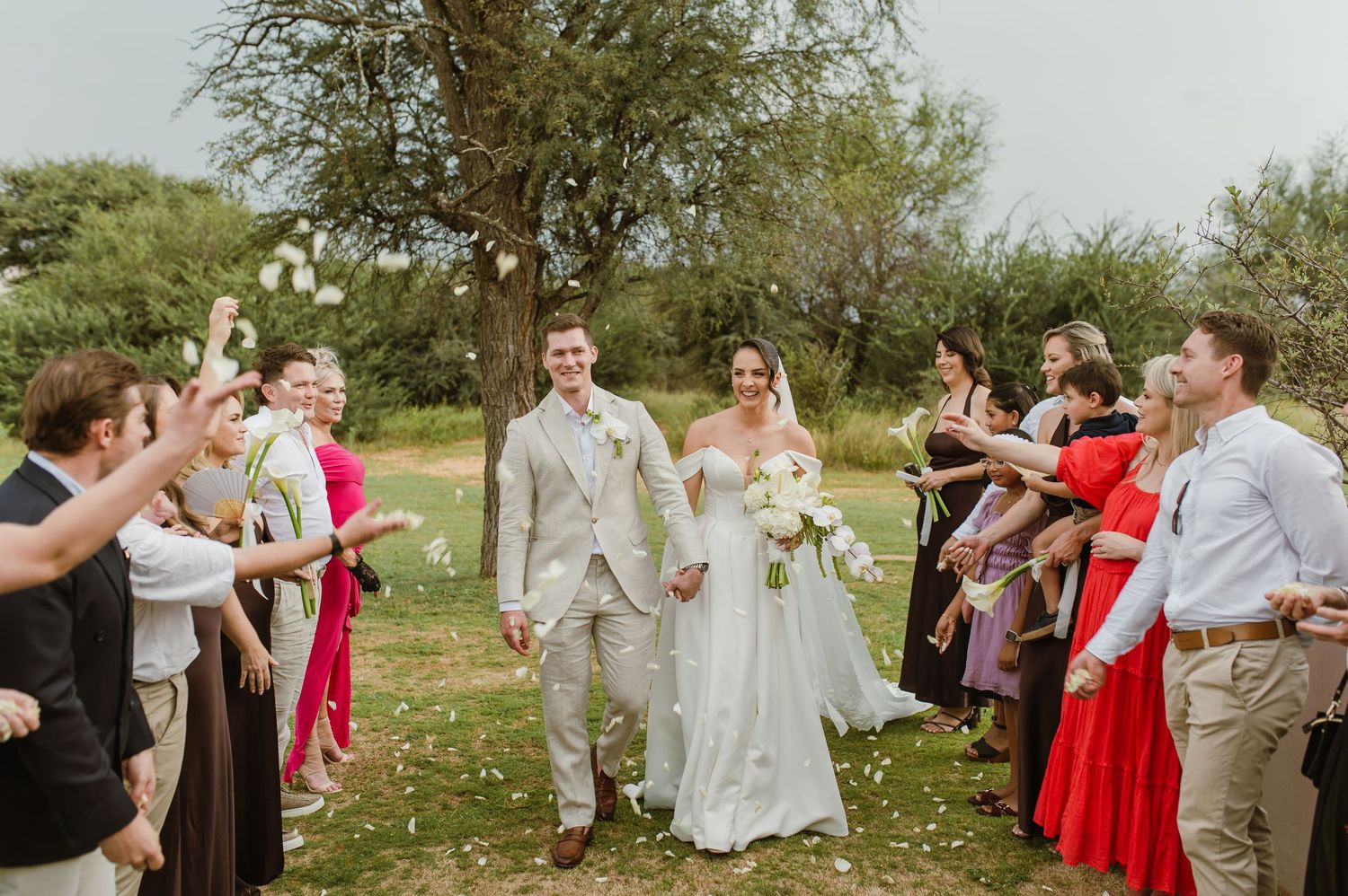 Wedding party and guests form a celebratory line outdoors under trees.