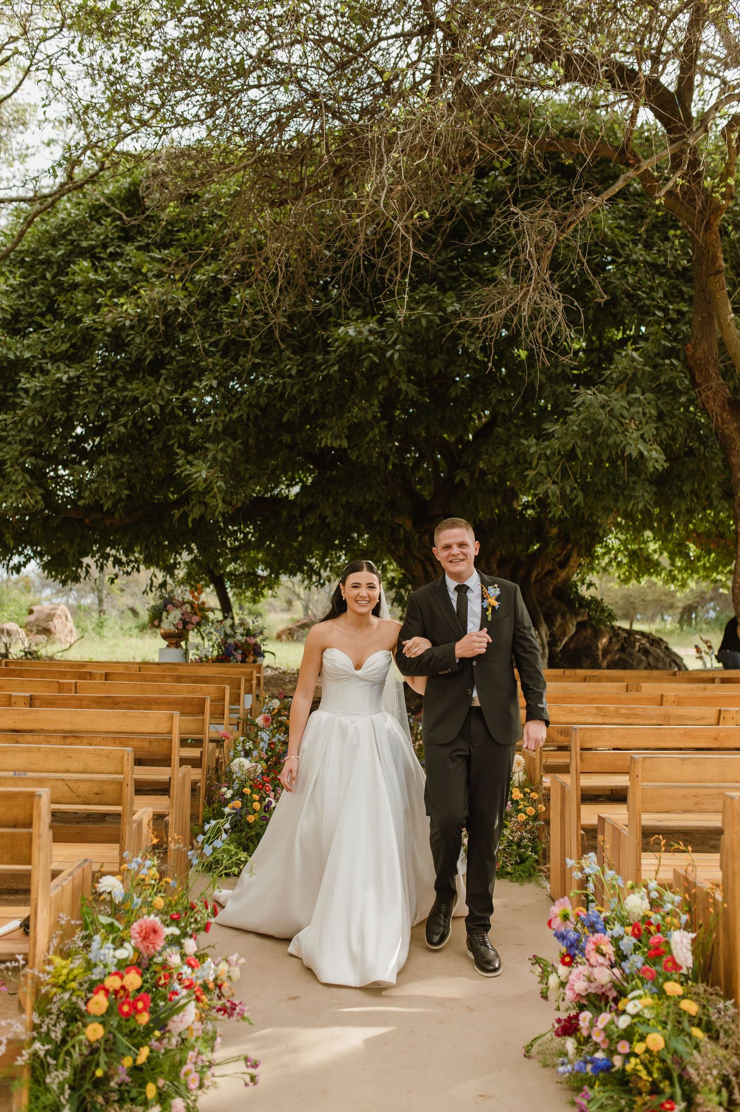 Newlyweds walking down floral-lined aisle after ceremony under a large tree outdoors.