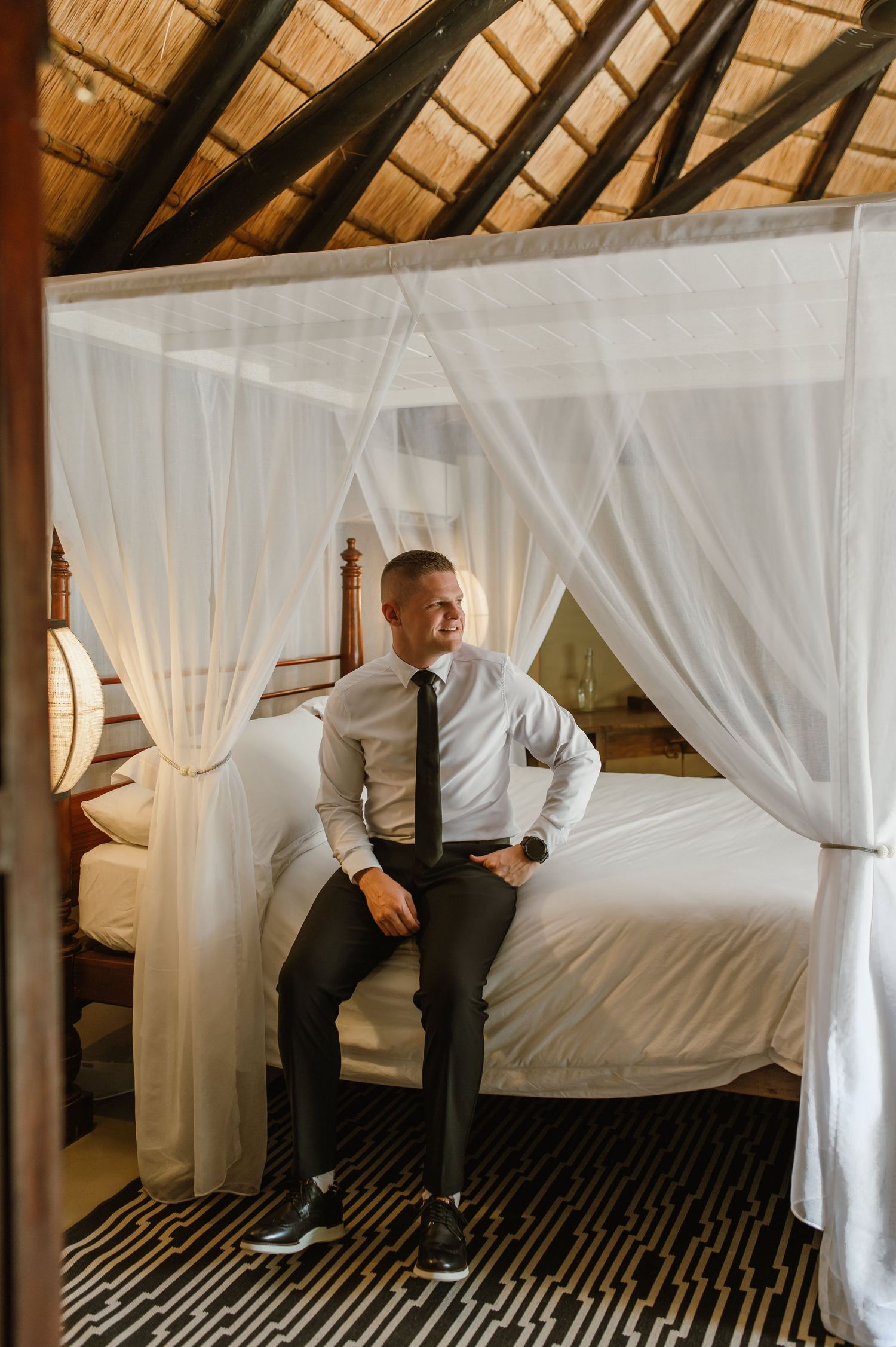 Groom in white shirt and black tie sitting on a canopy bed in a luxury safari lodge room.