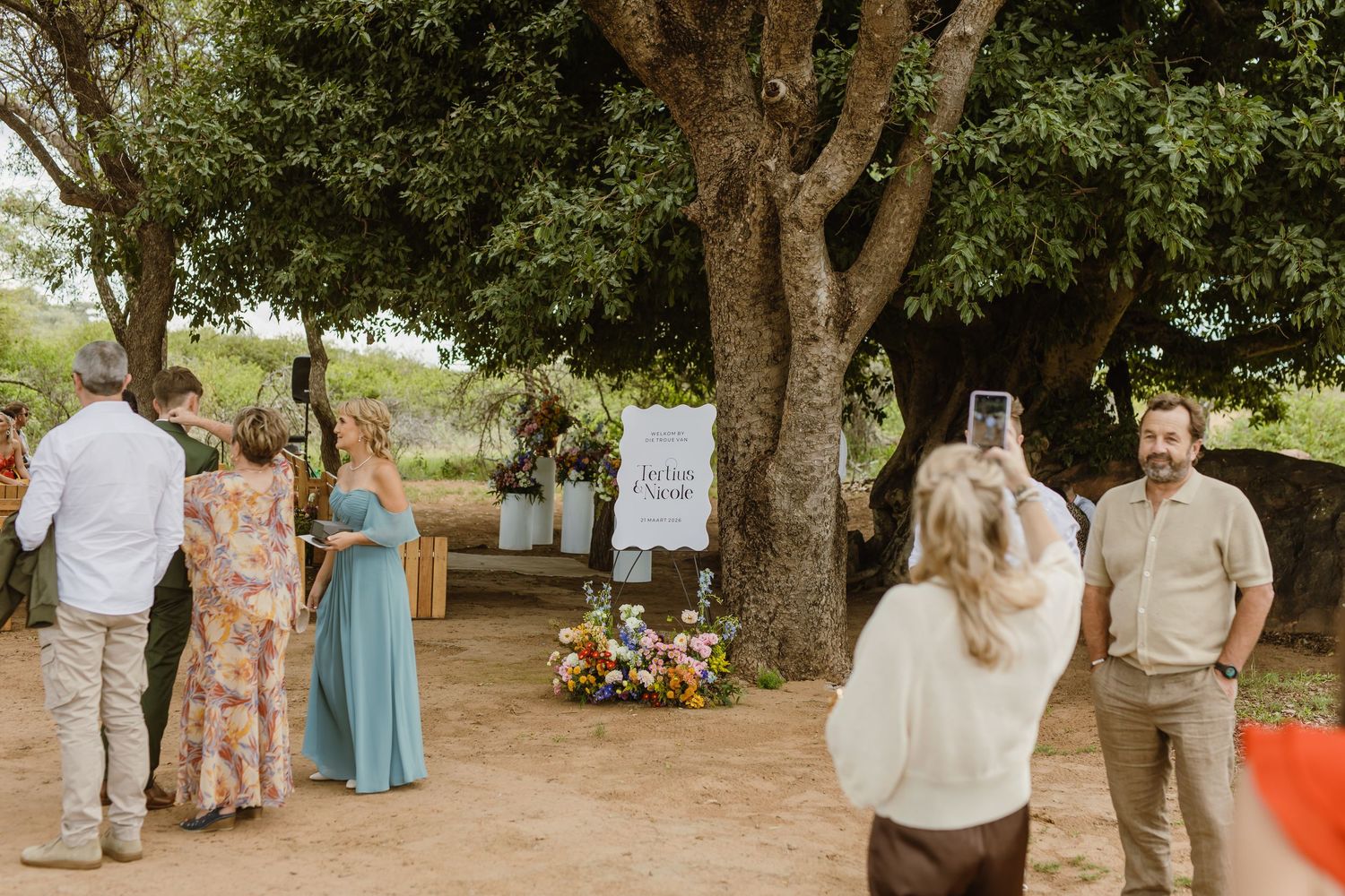 Wedding guests gathering outdoors near a large tree with floral arrangements and signage.