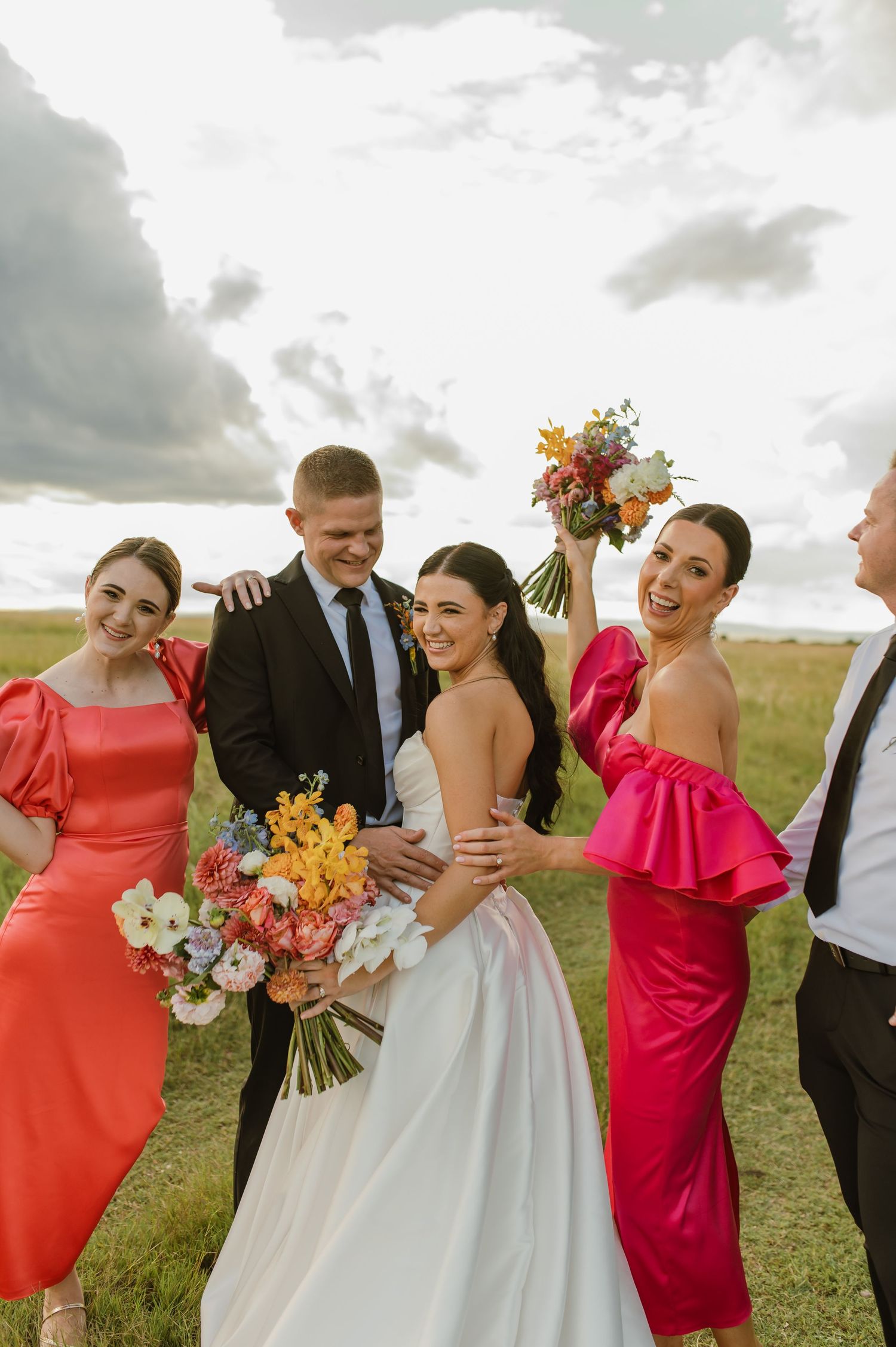 Wedding party laughing together outdoors, bride holding colorful bouquet with bridesmaids in bright dresses.