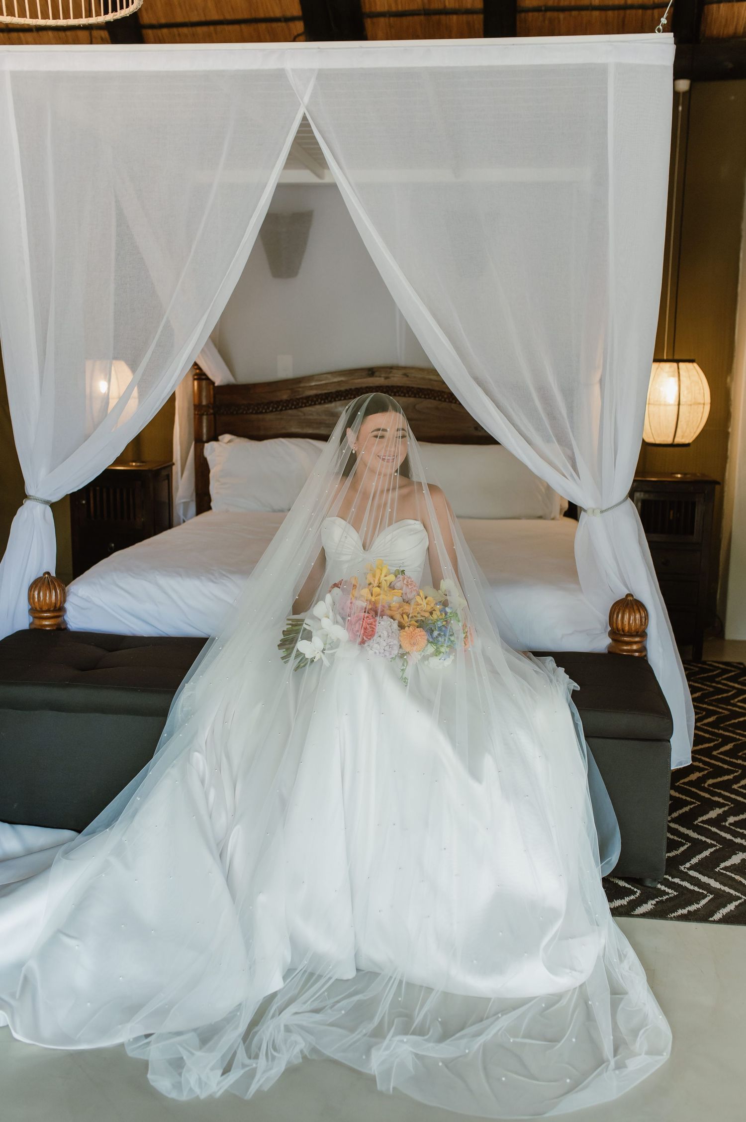 Bride in a full white ball gown and veil sitting in front of a four-poster canopy bed holding a floral bouquet.