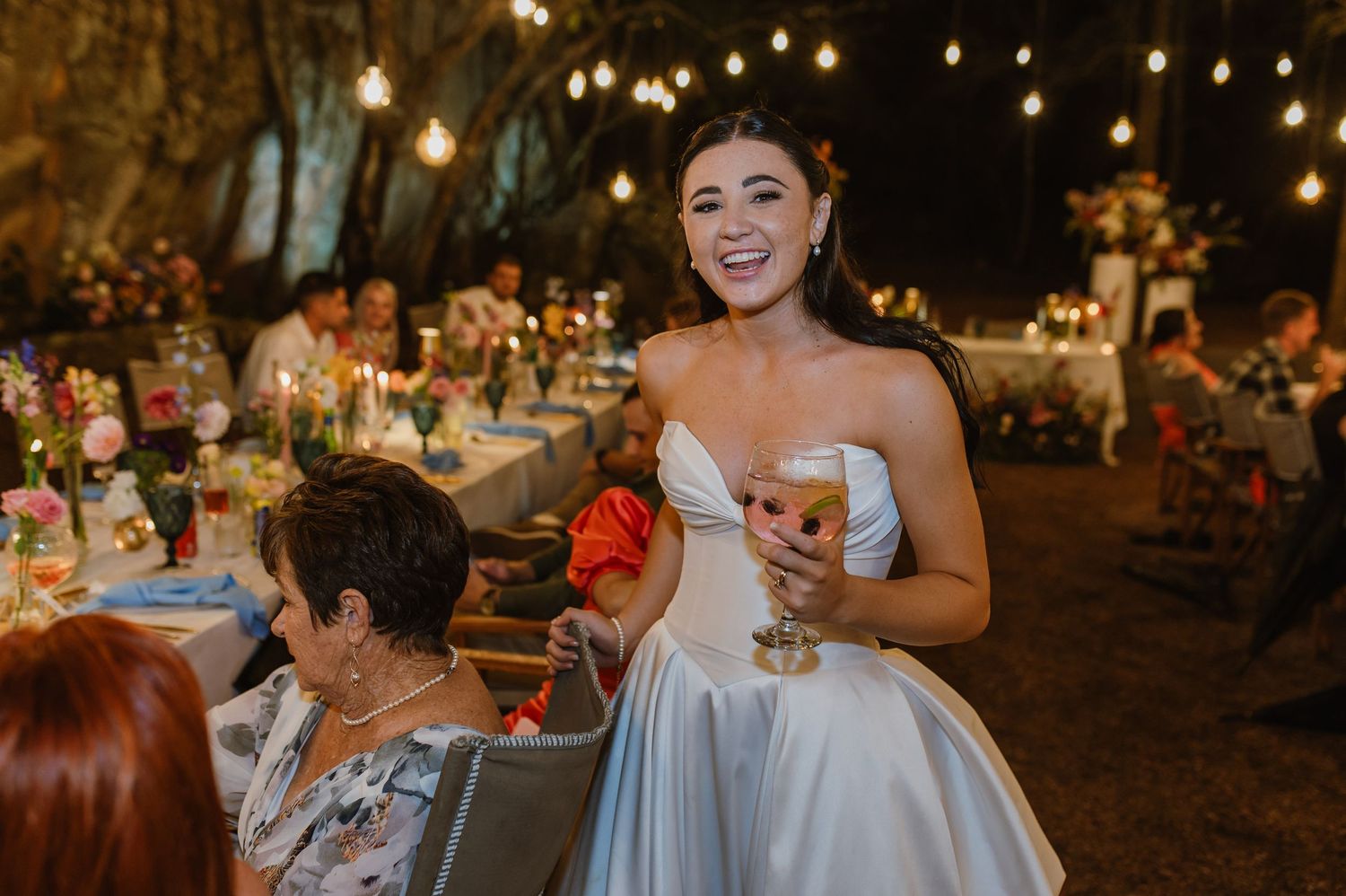 Radiant bride in strapless gown holding a drink and smiling at outdoor wedding reception with string lights.