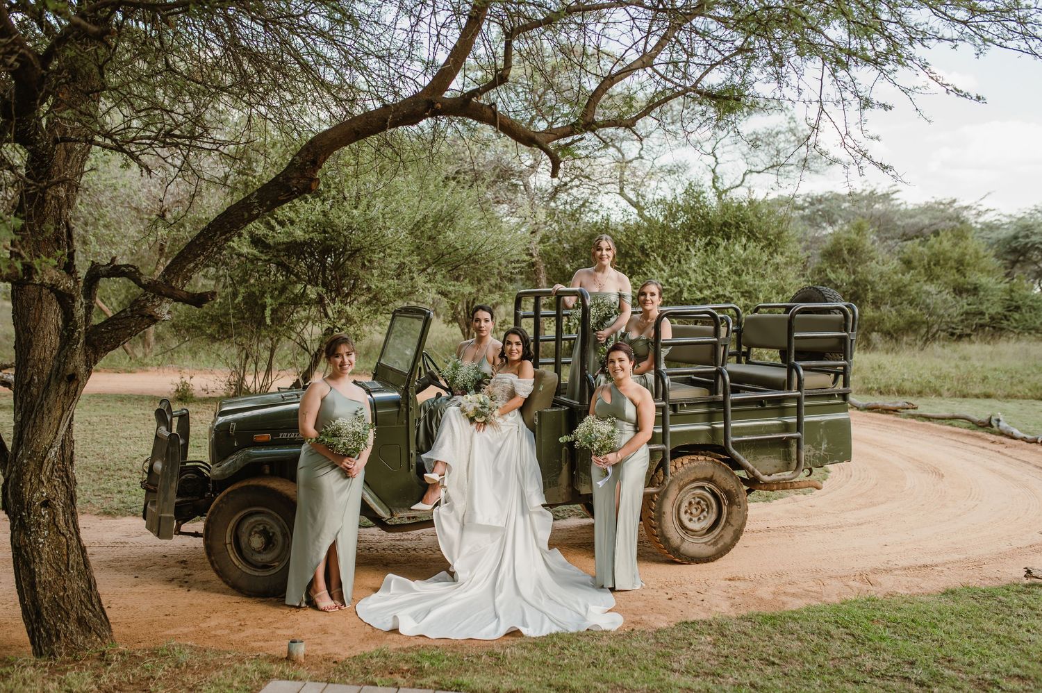Wedding party gathered around vintage military vehicles under shady trees on a dusty safari road.