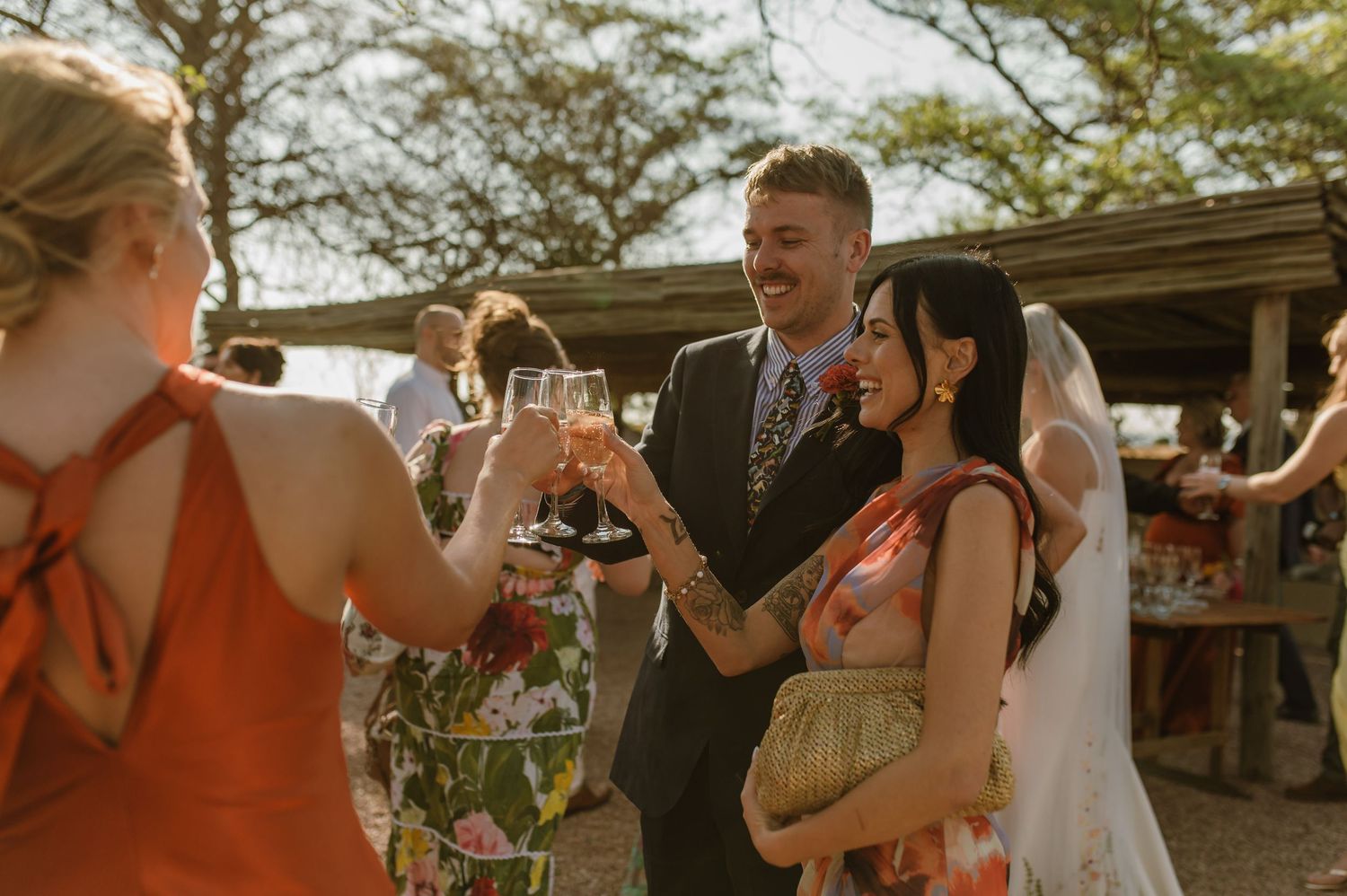 Wedding guests celebrate outdoors at sunset with drinks and laughter at a rustic venue.