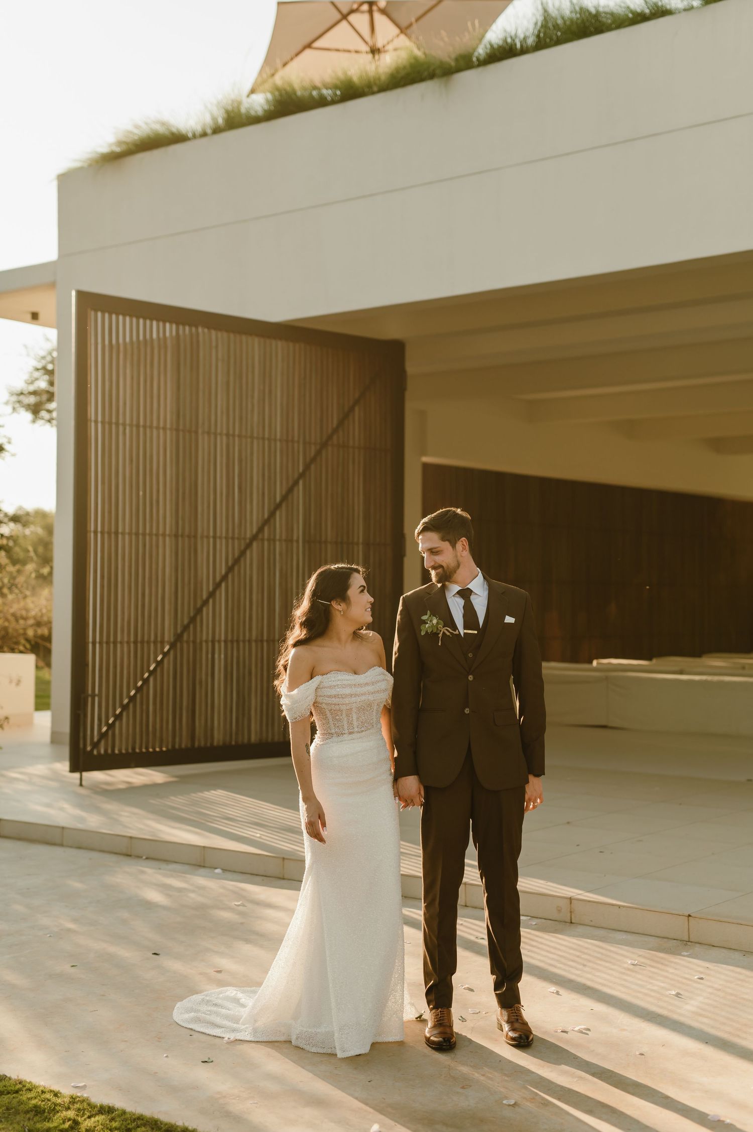 Elegant couple in wedding attire poses against modern architectural backdrop at sunset.