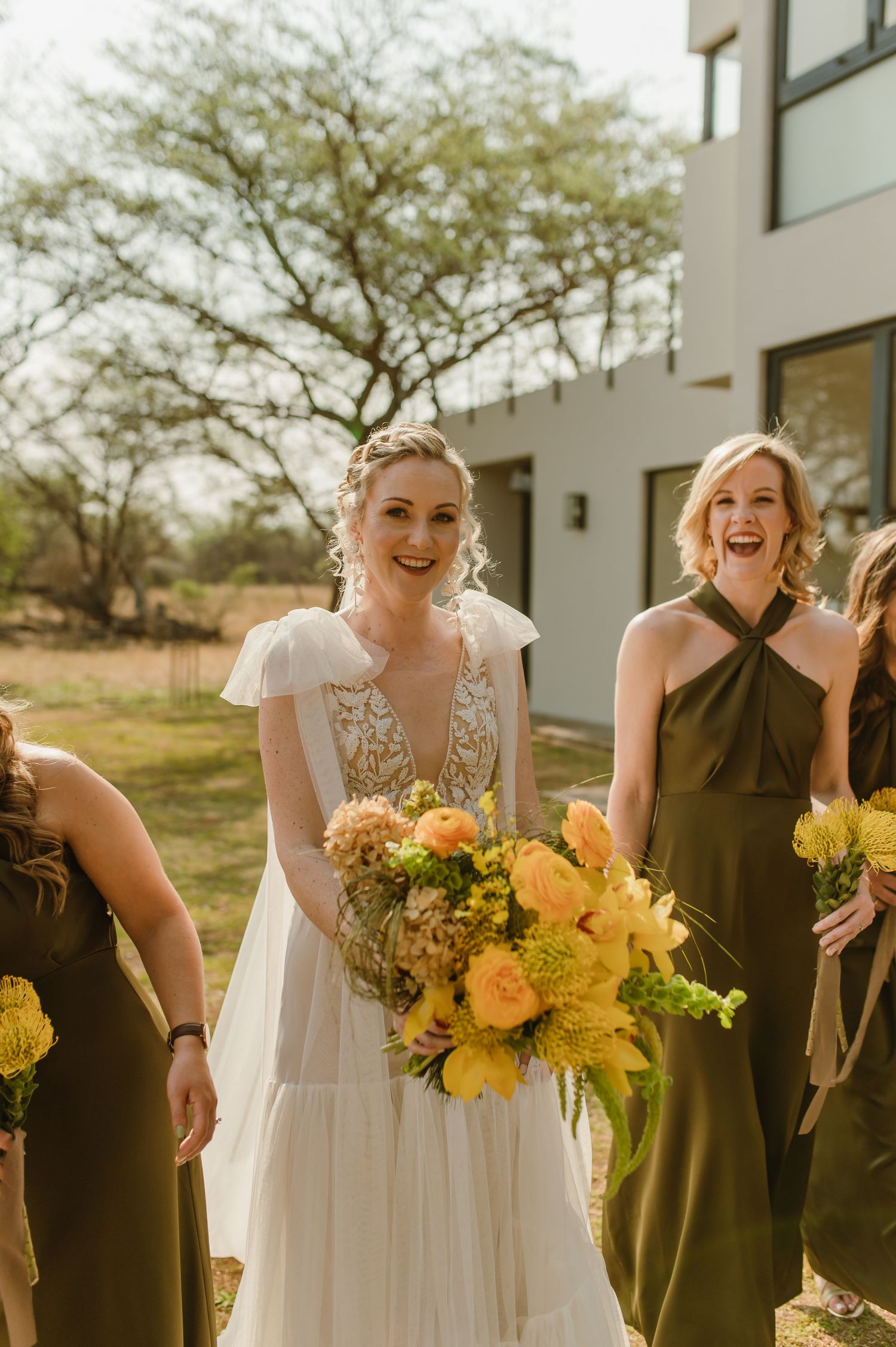 Bridesmaids in black dresses holding yellow sunflower bouquets laugh together outdoors.