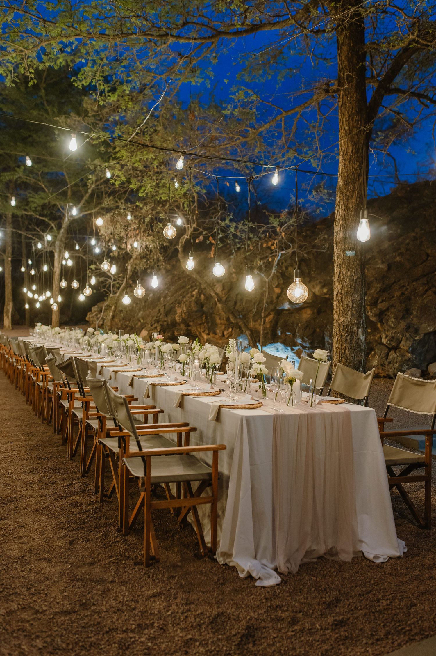 Outdoor wedding reception setup with string lights hanging between trees and white-clothed tables.