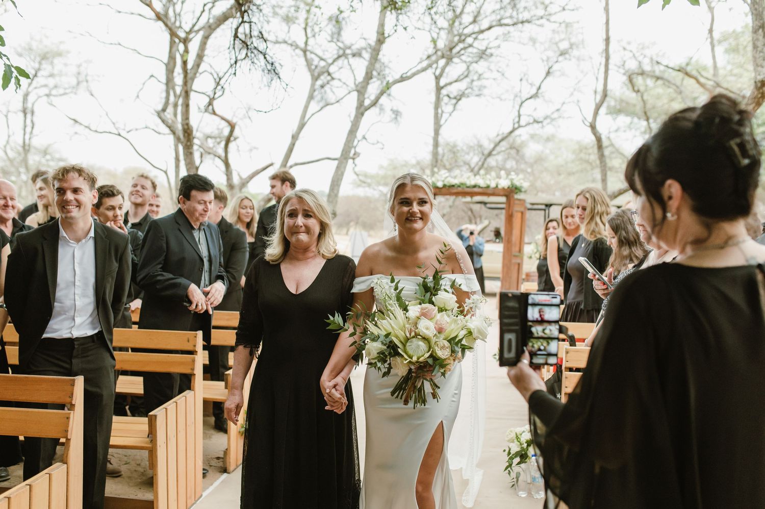 Wedding party and guests gathered for an outdoor ceremony with natural woodland backdrop.