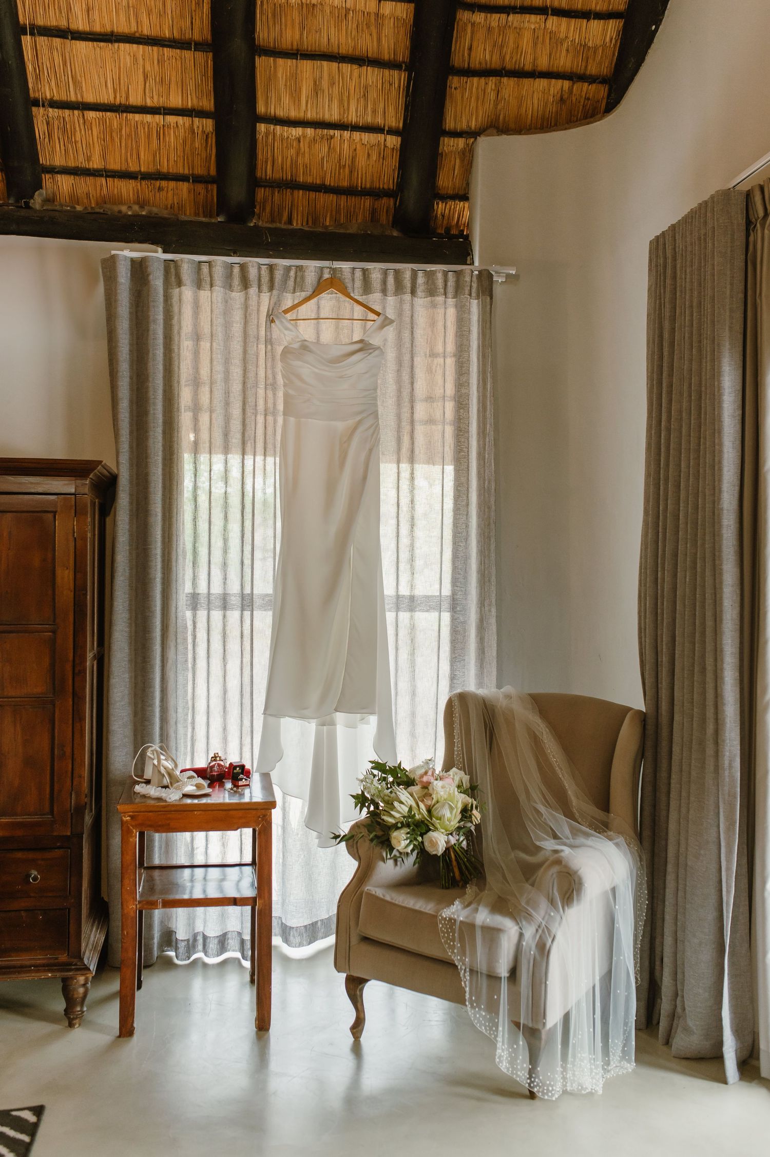 White wedding dress hanging in room with thatched roof and elegant armchair.