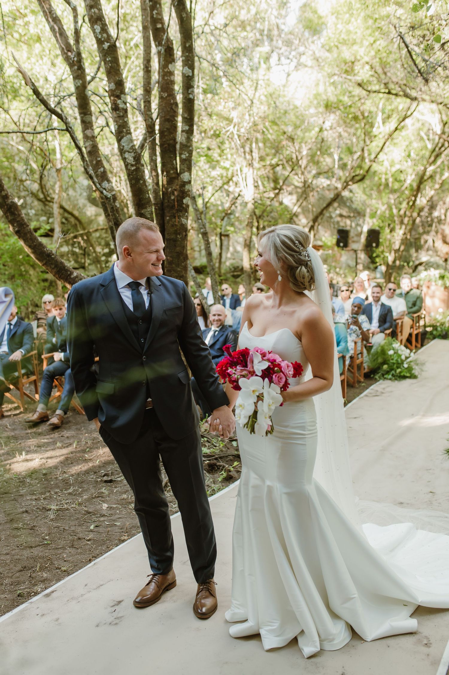 A couple exchanges vows during their outdoor woodland wedding ceremony on a sunlit path.