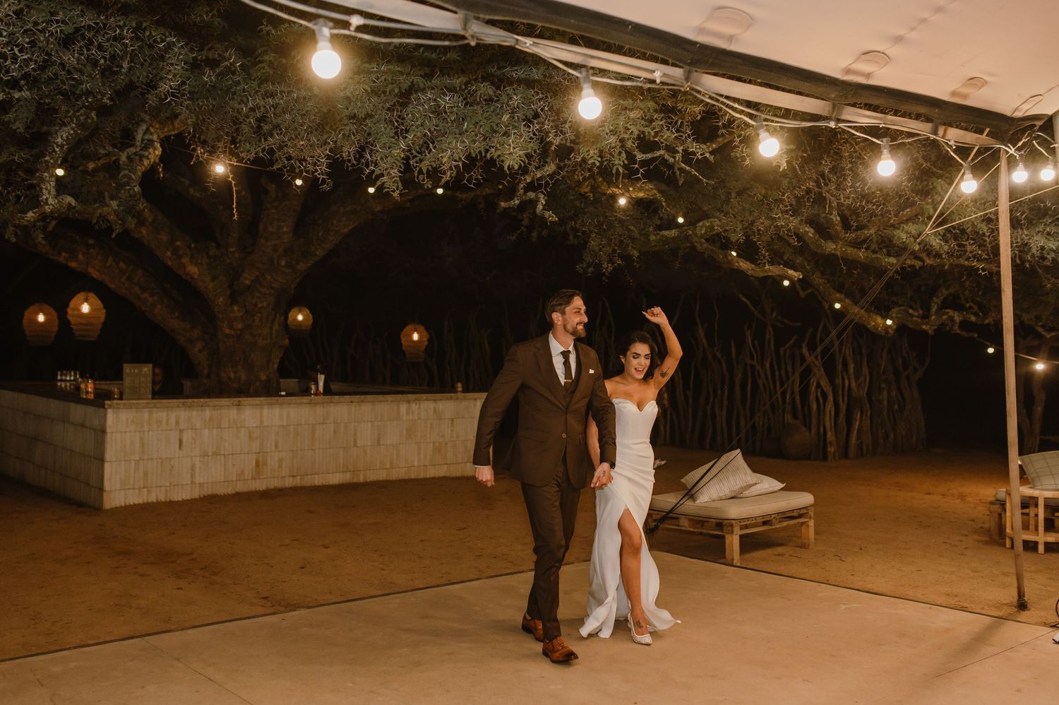 String lights illuminate an outdoor wedding reception space under a tent at twilight.
