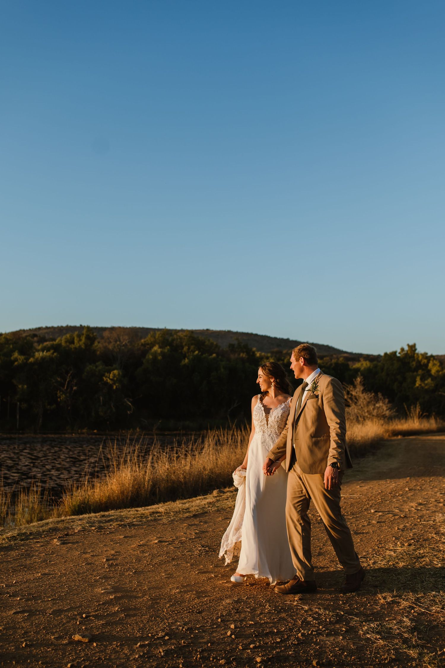 Newlyweds walk together on a dirt path during sunset with mountains in the background.