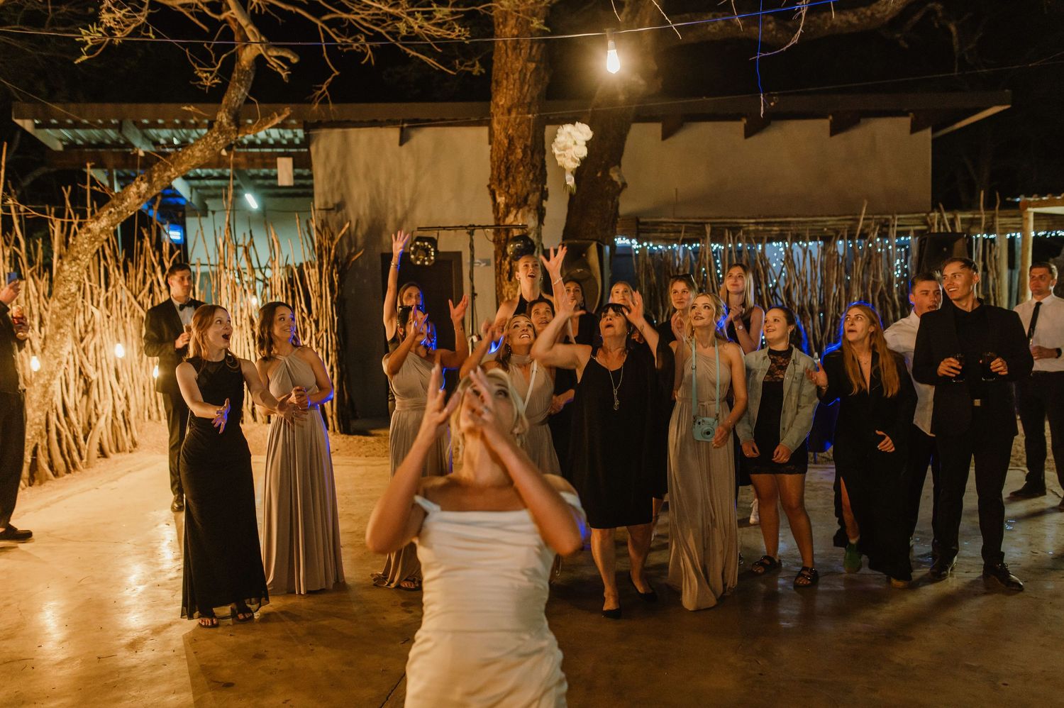 Large group celebrates at rustic indoor-outdoor venue with dramatic lighting and natural decor.