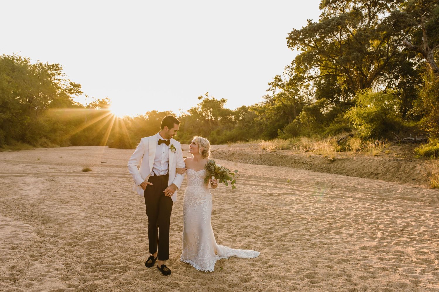 Wedding couple poses together on a sandy beach during golden hour with sunlight peeking through trees.