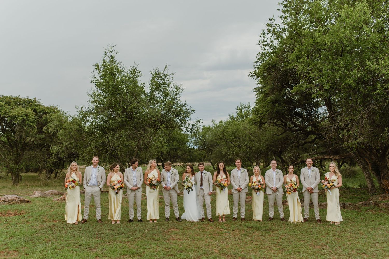 Large wedding party lined up in white and yellow attire for a panoramic photo in a natural setting.
