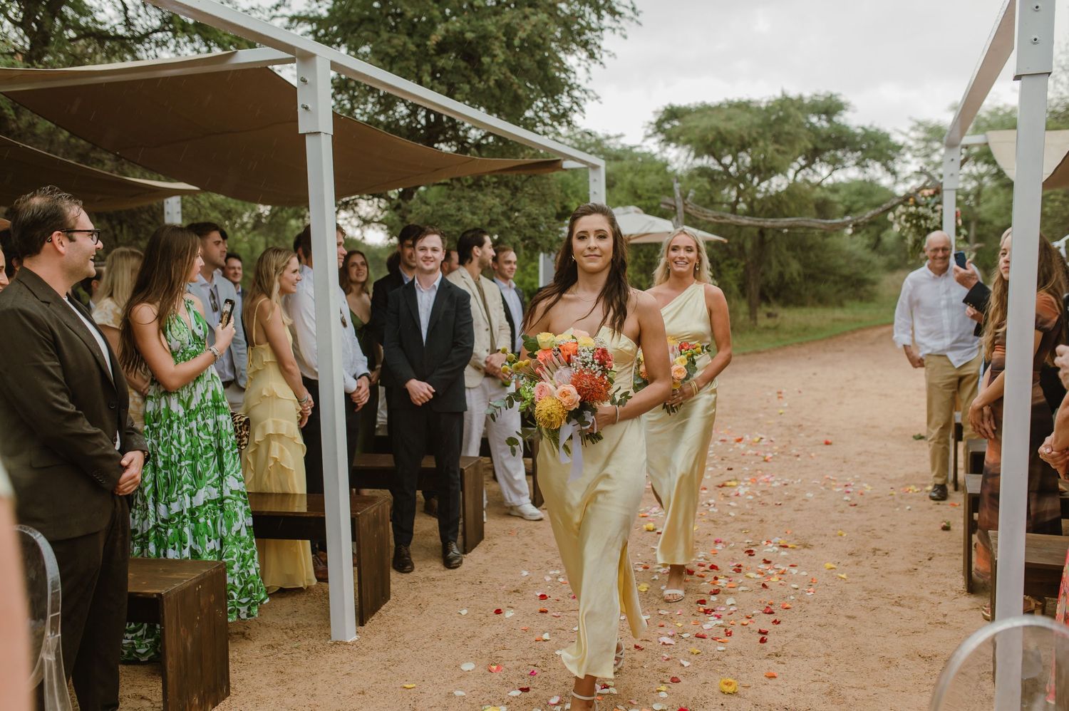 Wedding guests line a path with scattered rose petals under trees at an outdoor ceremony.