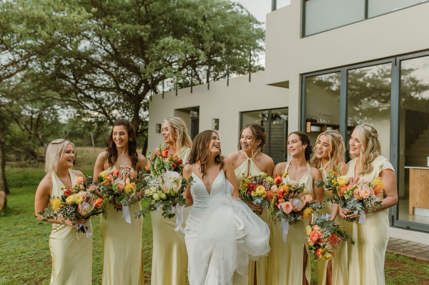 Bridal party in yellow dresses standing outdoors with floral bouquets.
