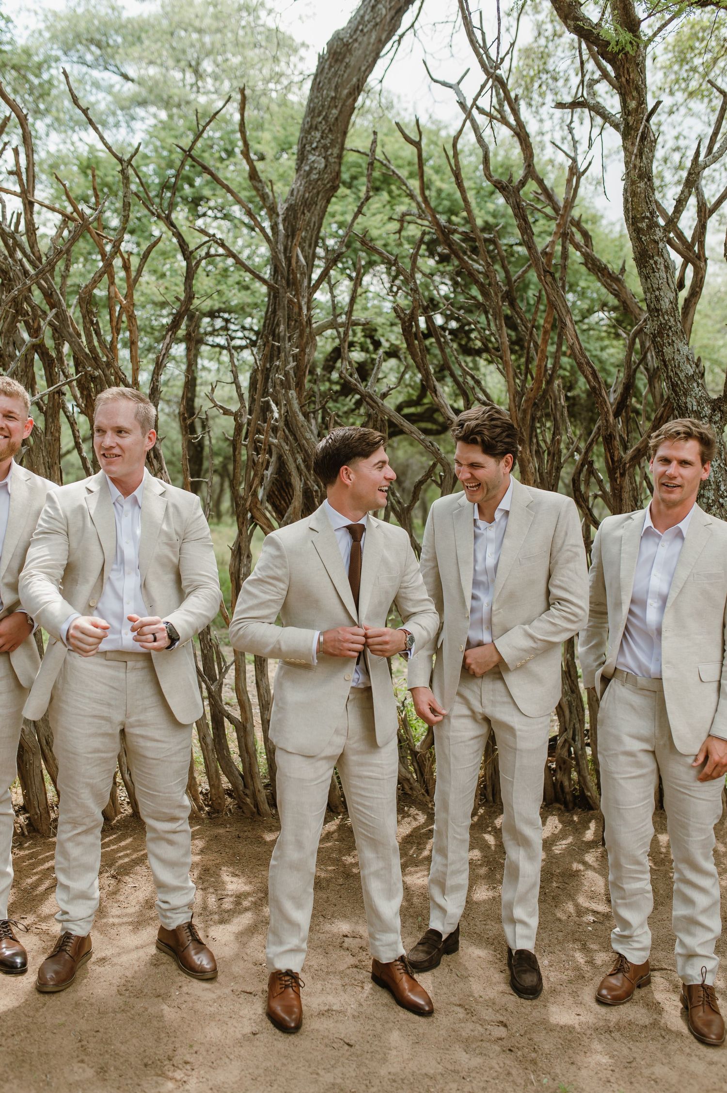 Groomsmen in beige suits and brown shoes stand in a casual line formation among trees.