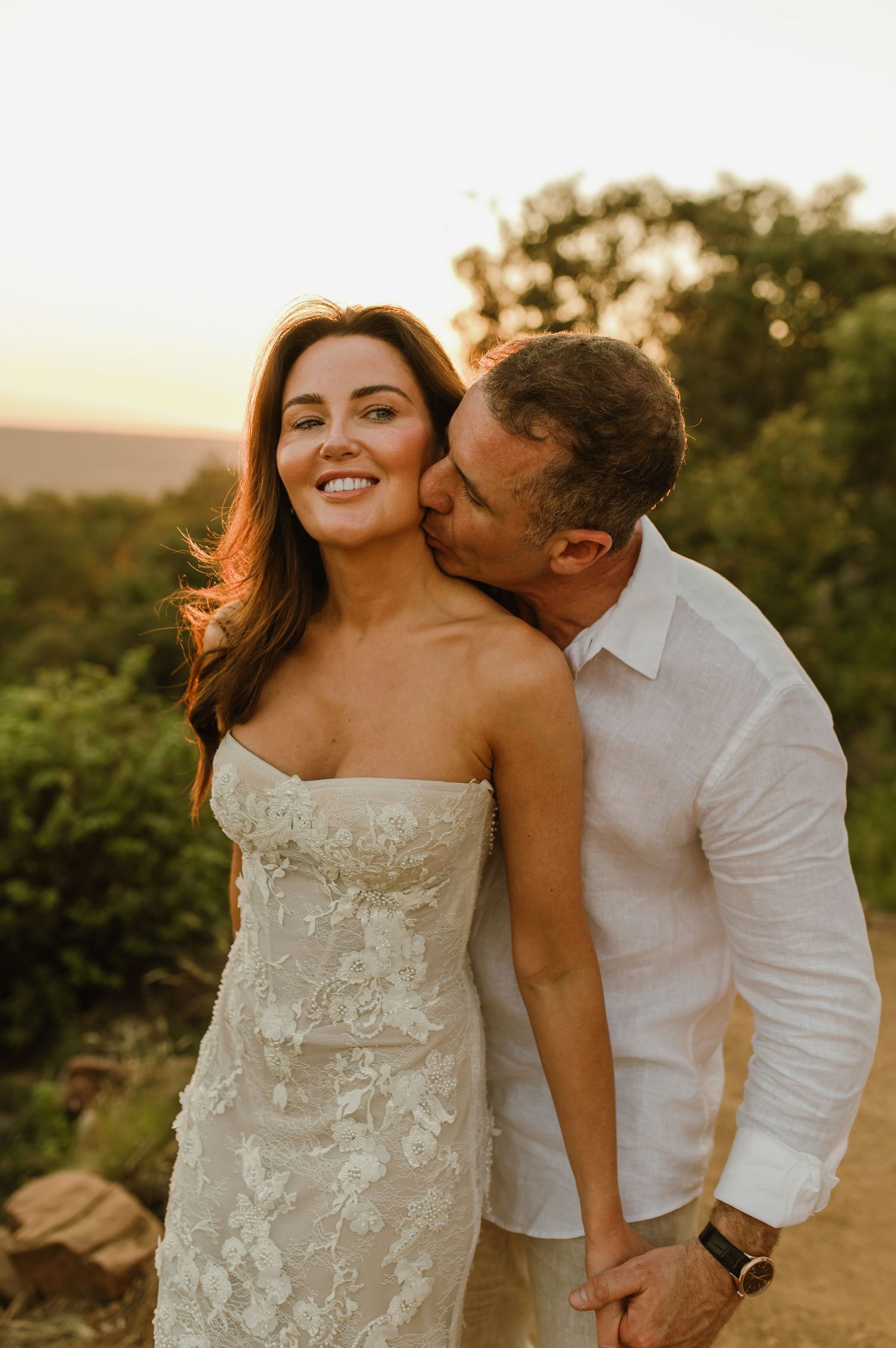 Groom kissing bride on the cheek as she smiles during an outdoor sunset wedding portrait.