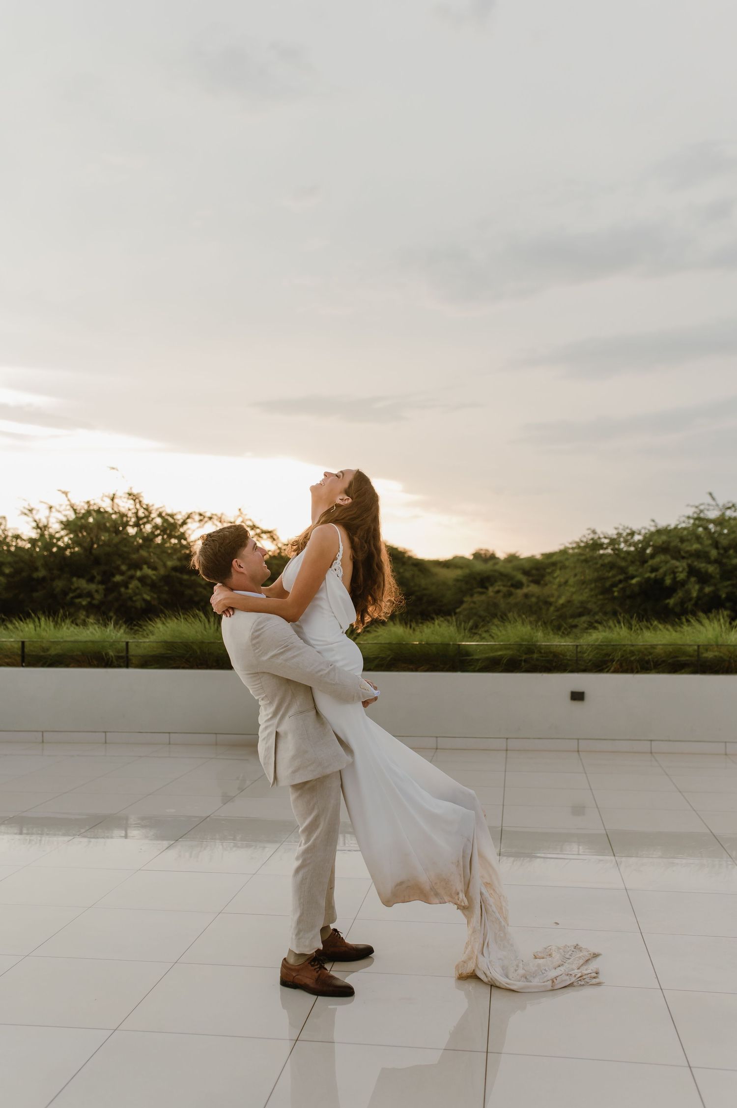 A couple in wedding attire share a joyful dance pose on an outdoor terrace at sunset.