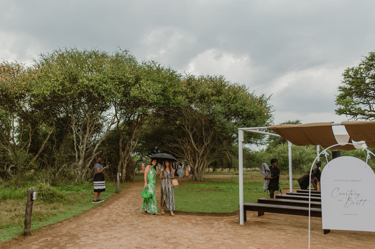 Outdoor wedding ceremony setup with wooden benches and natural decor under trees on a dirt path.
