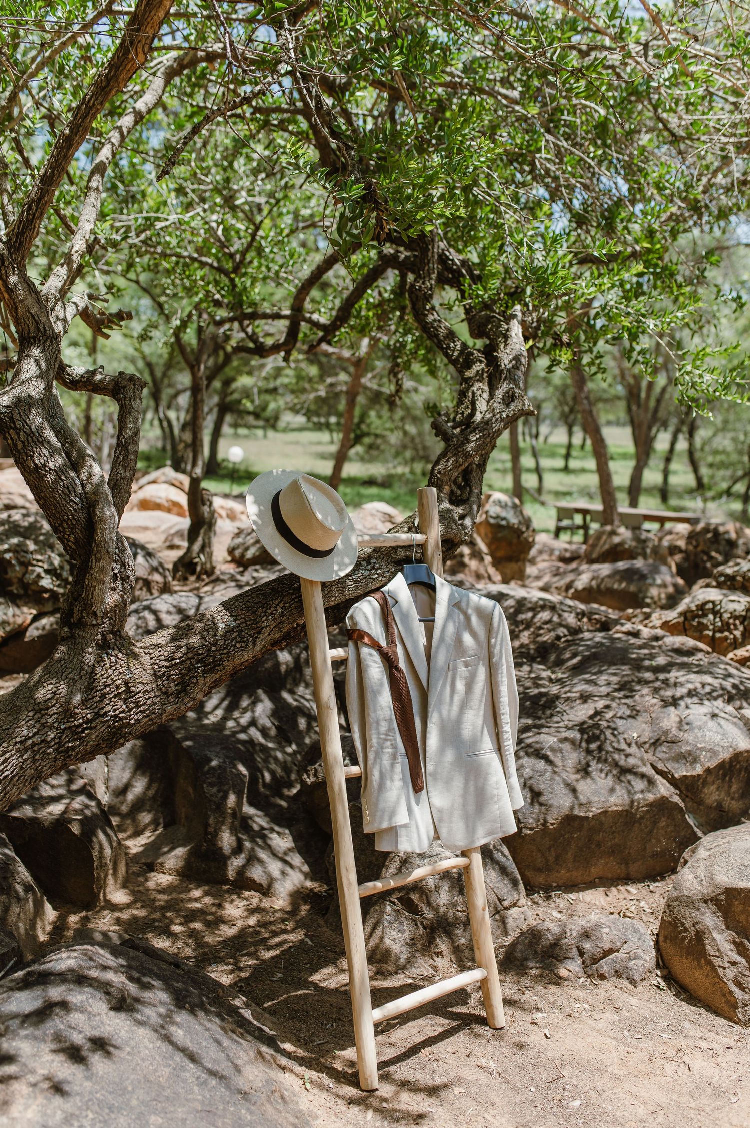 Wedding attire hanging on wooden stands outdoors under shady trees.