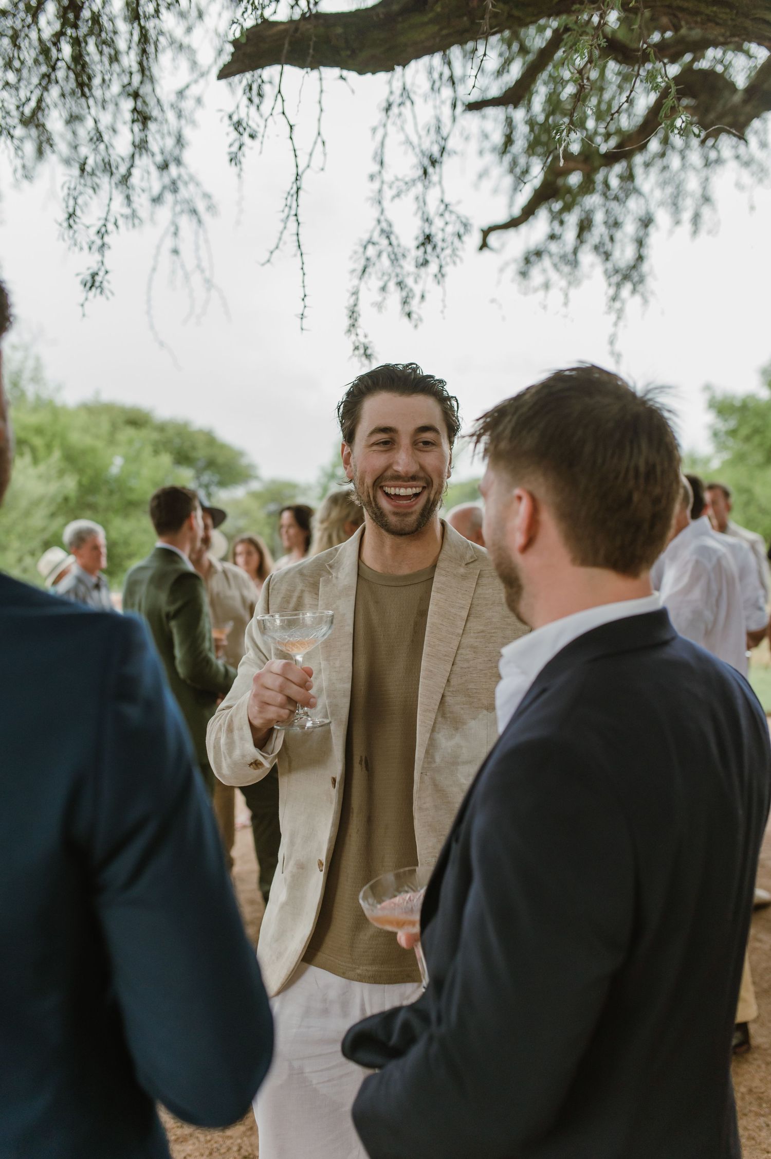 Wedding guests share laughter and drinks during an outdoor reception under trees.