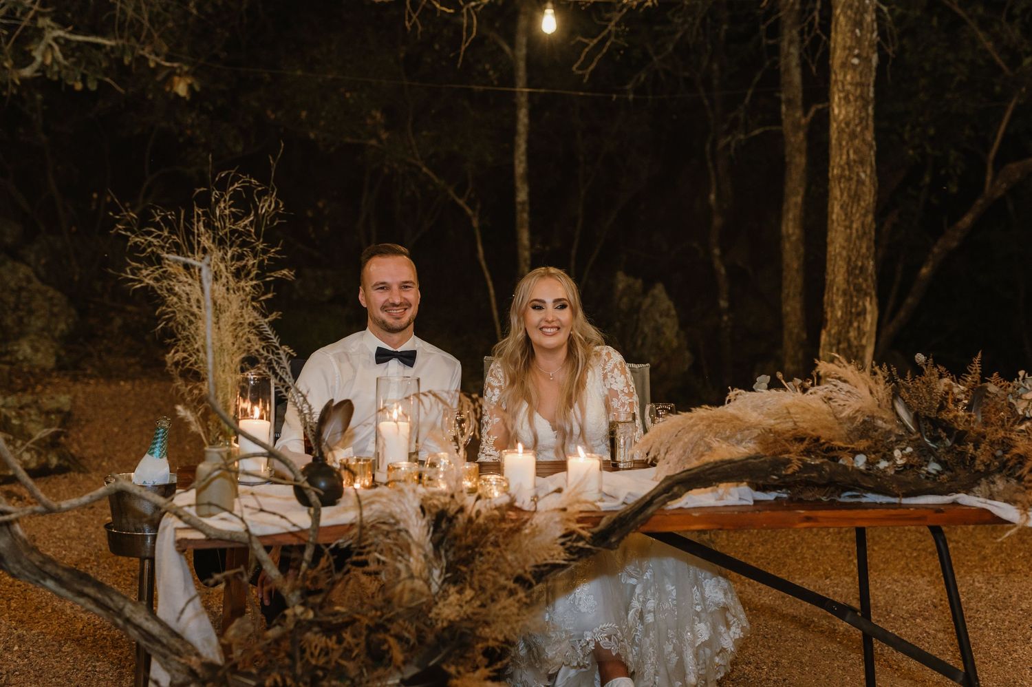 Couple seated at elegantly decorated outdoor reception table with candlelight and rustic decor at night.
