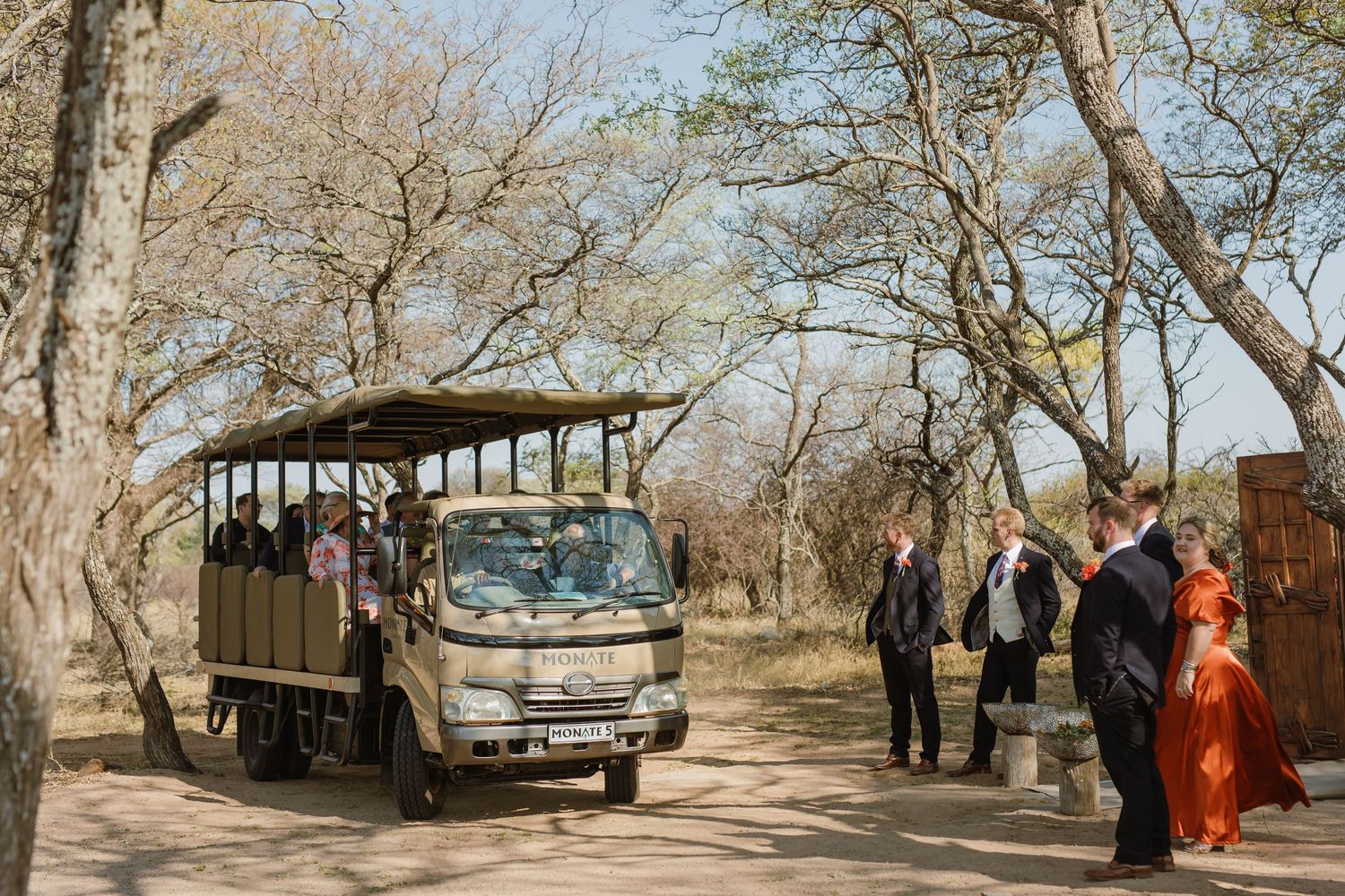 Safari vehicles and wedding party gathered under acacia trees in natural bushveld setting.
