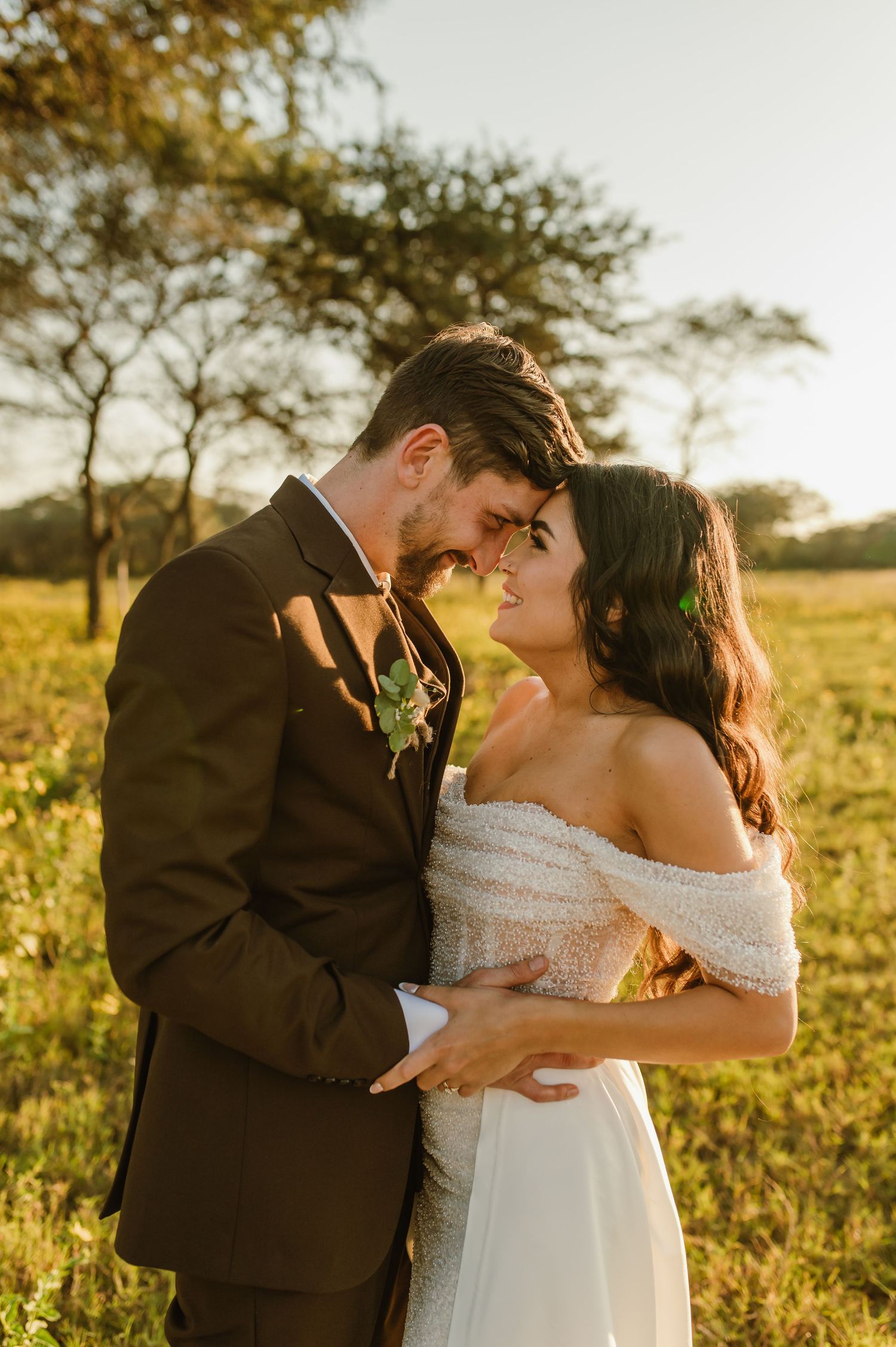 A tender wedding moment captured in a sun-drenched field with acacia trees at sunset.