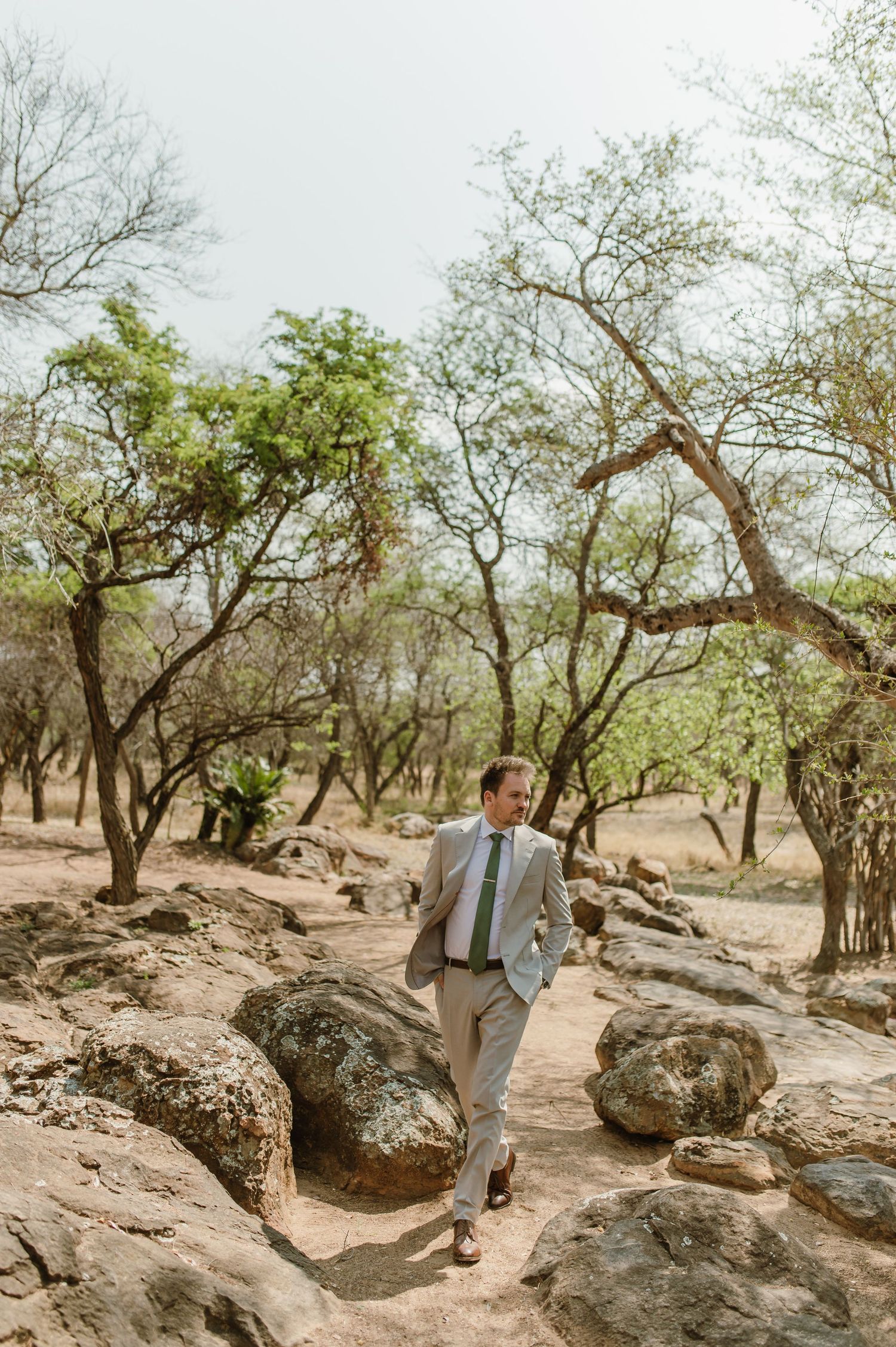 Person in formal attire walking among rocky terrain and trees.